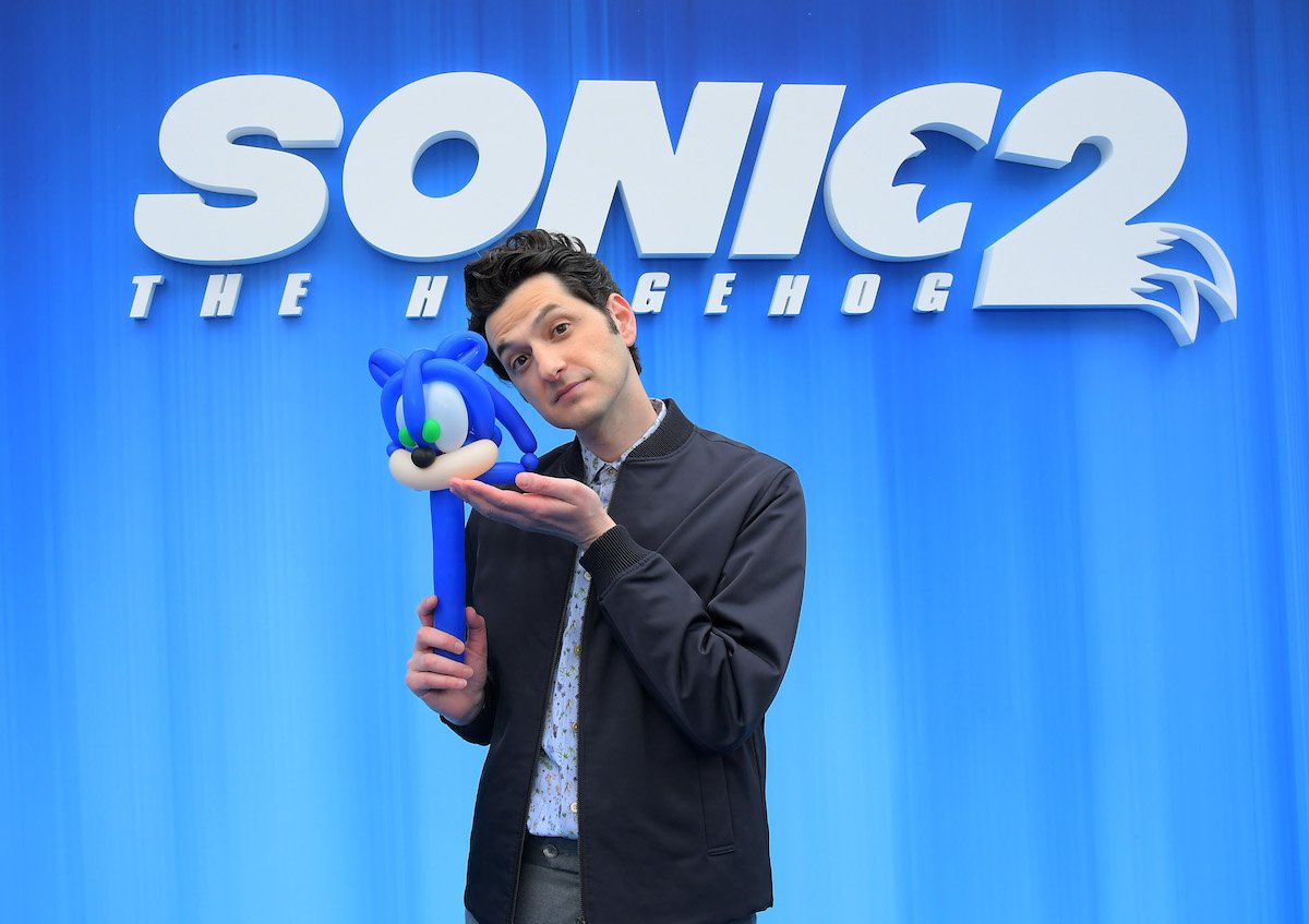 Sonic the Hedgehog 2 actor Ben Schwartz in front of a Sonic sign and holding a mini Sonic
