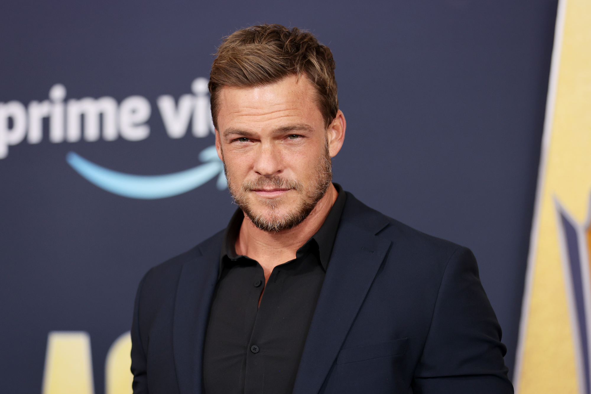 Alan Ritchson wearing a black suit in front of Prime Video step and repeat