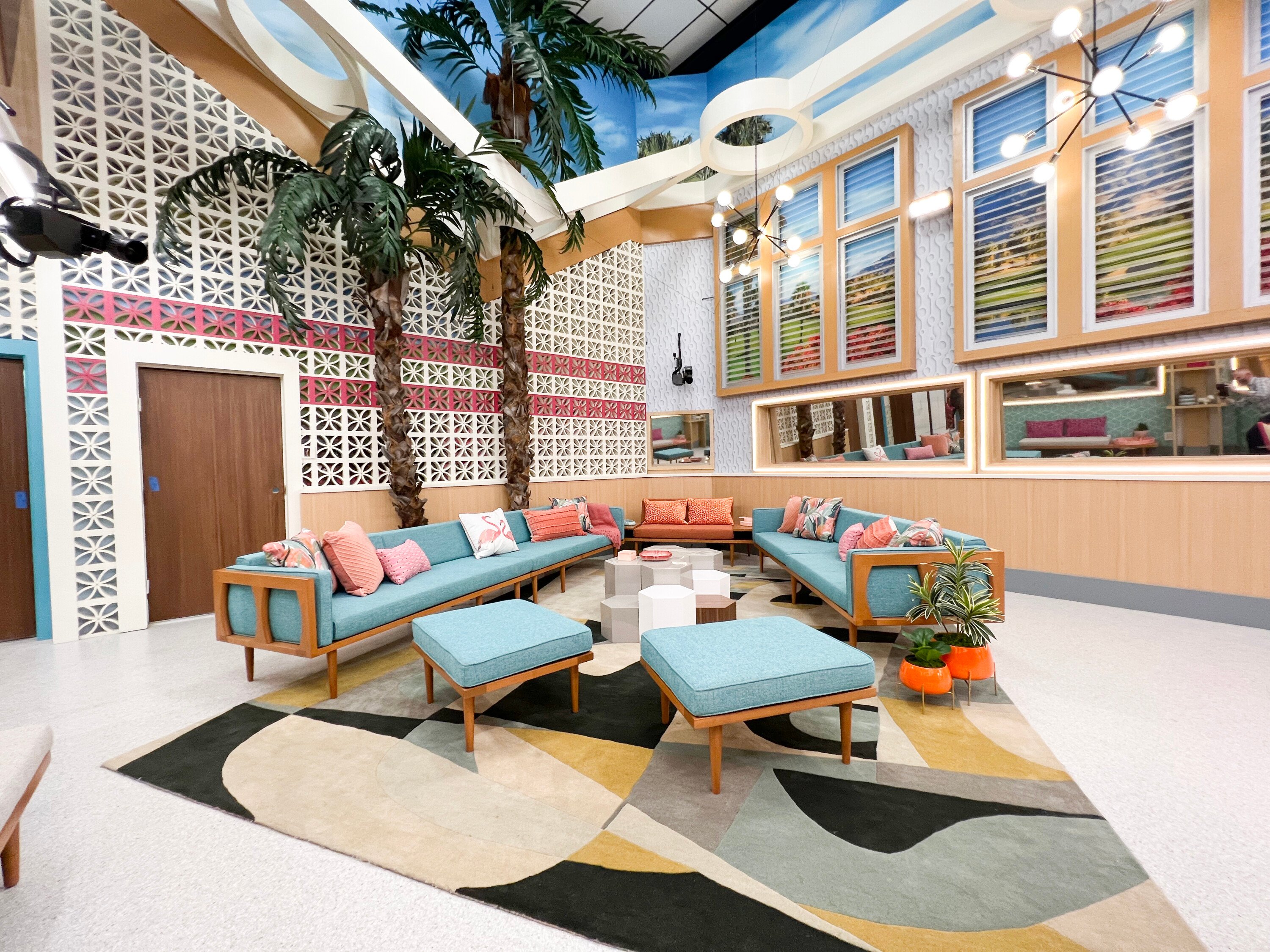 The living room of the 'Big Brother 24' house, which features fake palm trees, retro blue couches, and coral colored pillows.