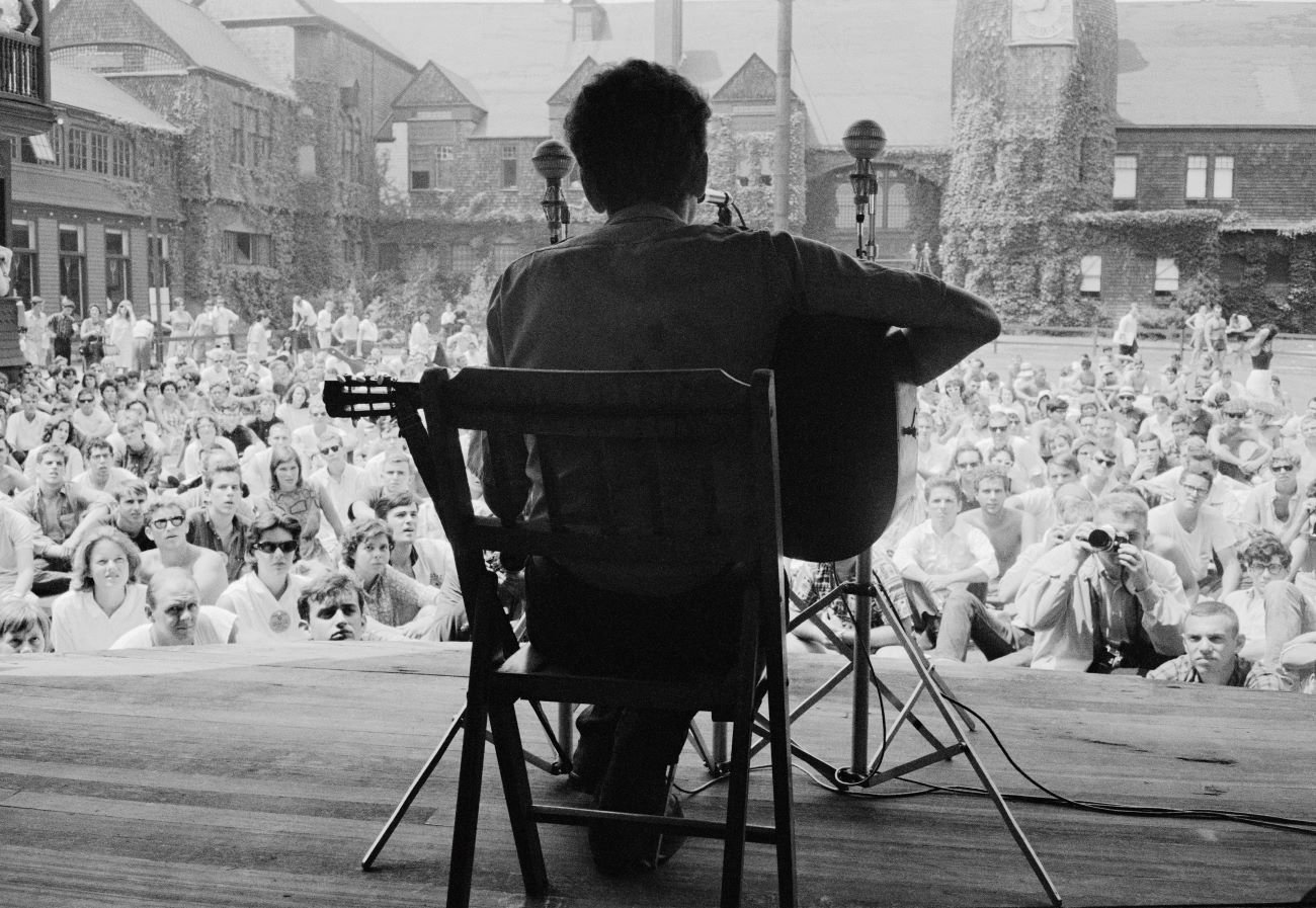 A black and white picture of Bob Dylan with his back to the camera. He sits in a chair and plays guitar for an audience.