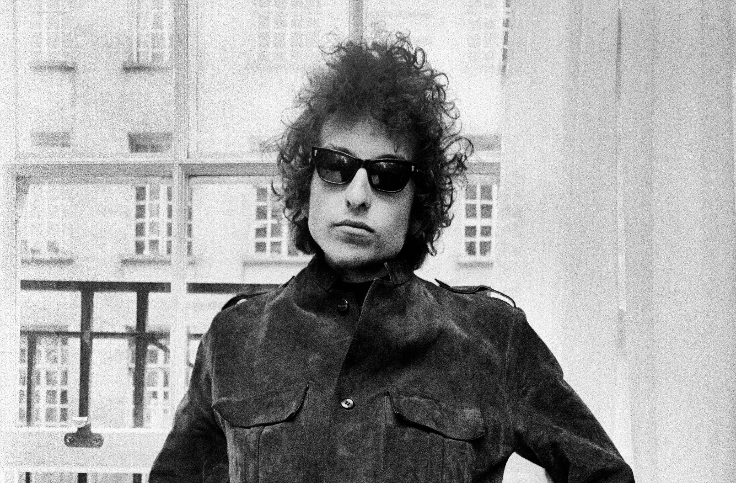 A black and white picture of Bob Dylan wearing sunglasses and sitting in front of a window. 