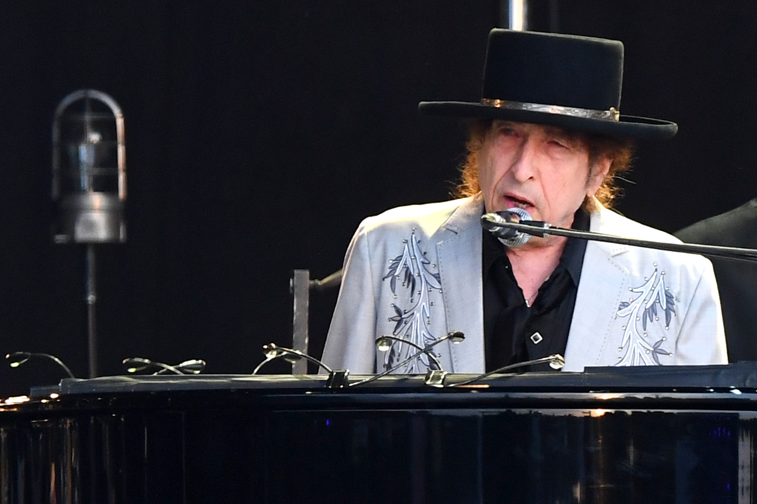 Bob Dylan sits at a piano and sings into a microphone at a venue in England.