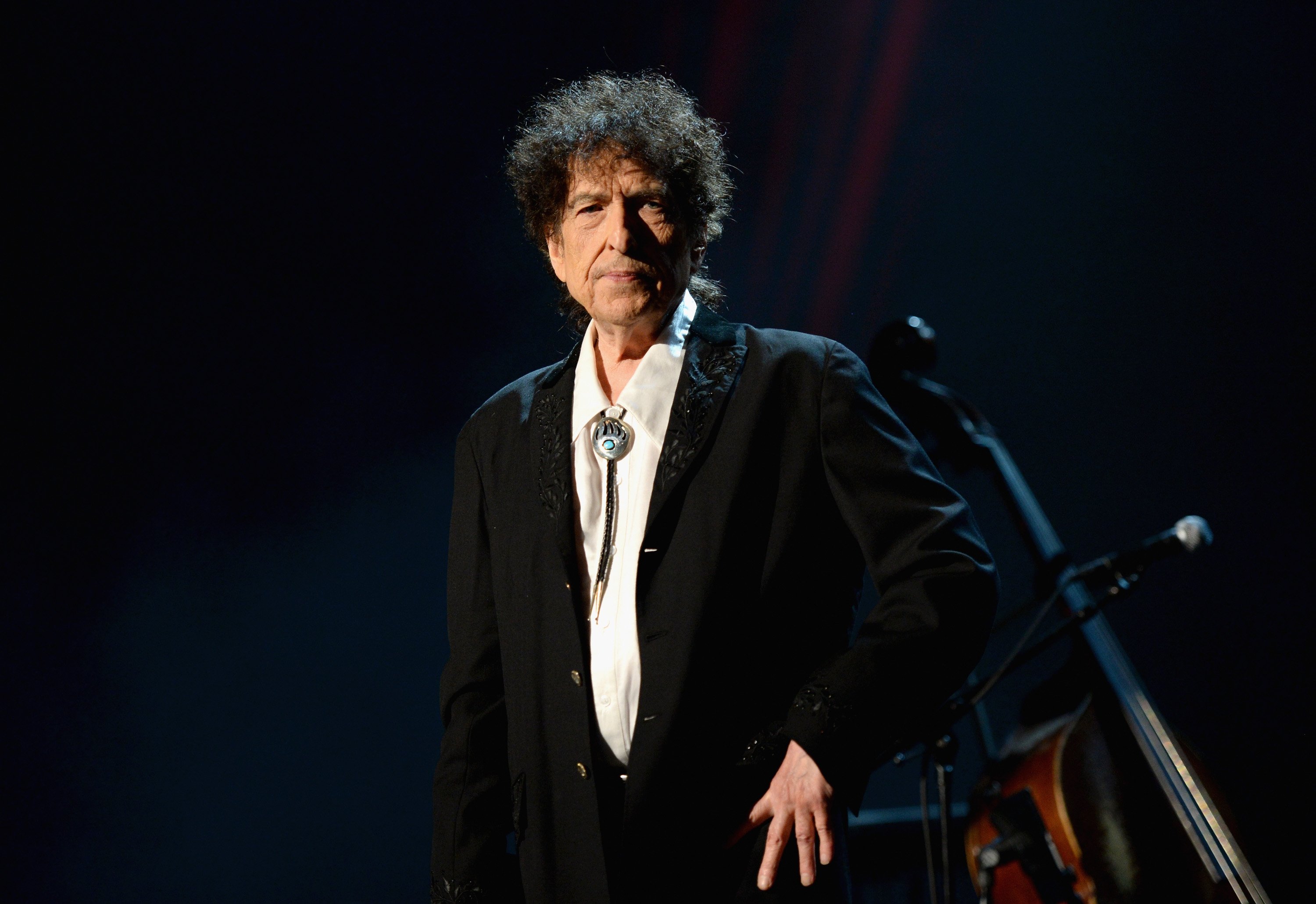 Bob Dylan wears a black jacket and a bolo tie and stands with his hand on his hip.