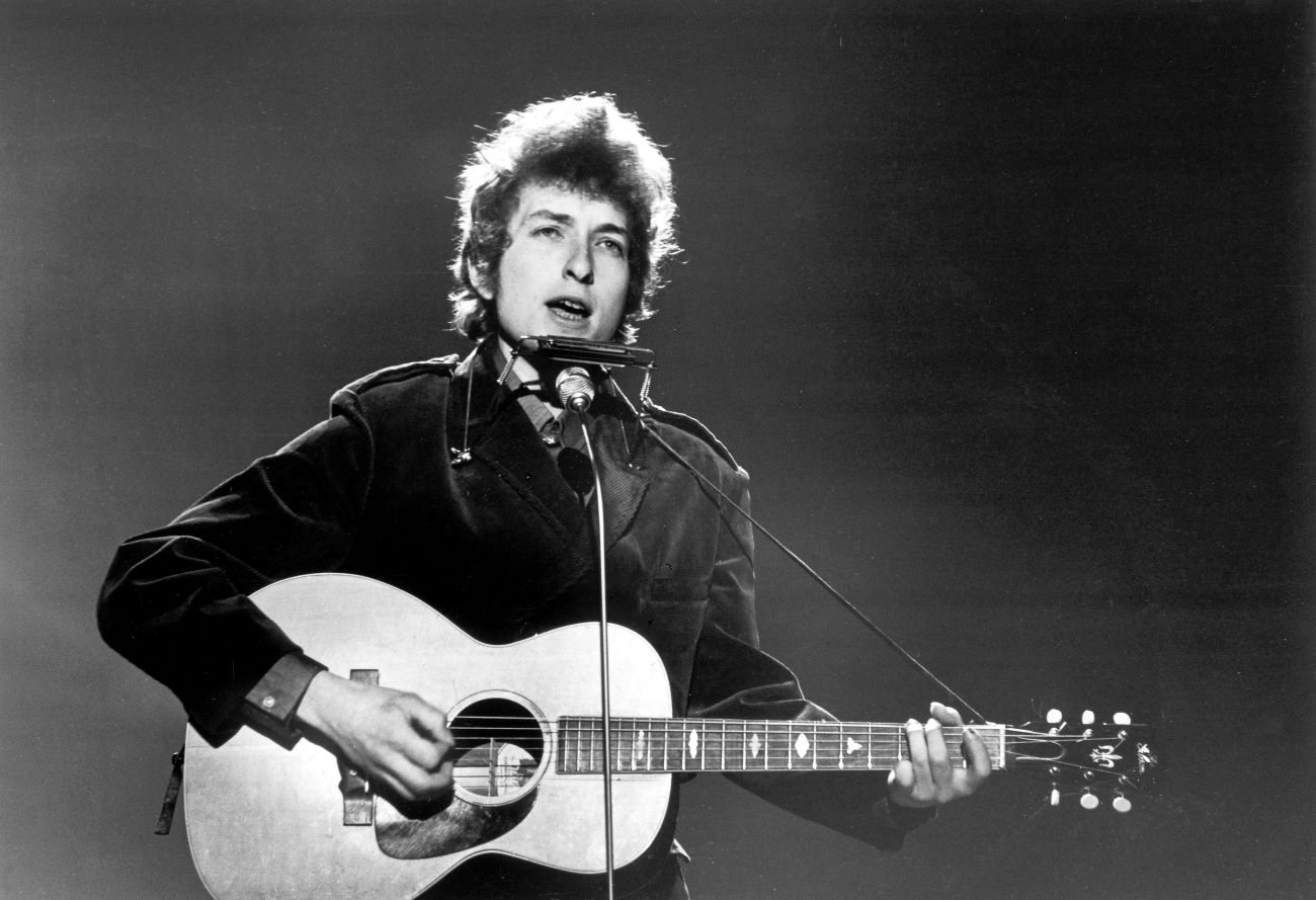 A black and white picture of Bob Dylan standing in front of a microphone with a guitar and harmonica.