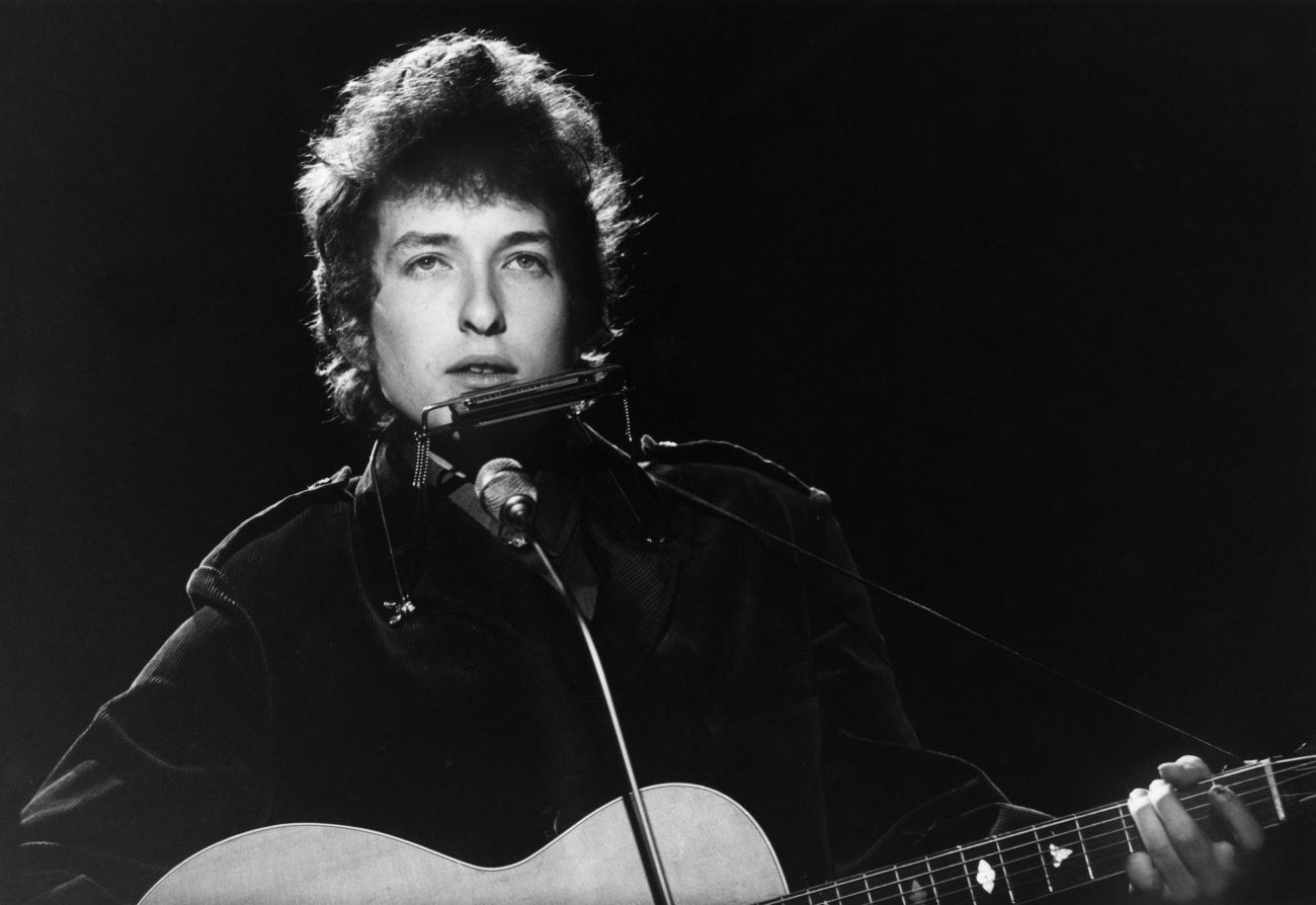 A black and white picture of Bob Dylan with a guitar and harmonica, standing in front of a microphone.
