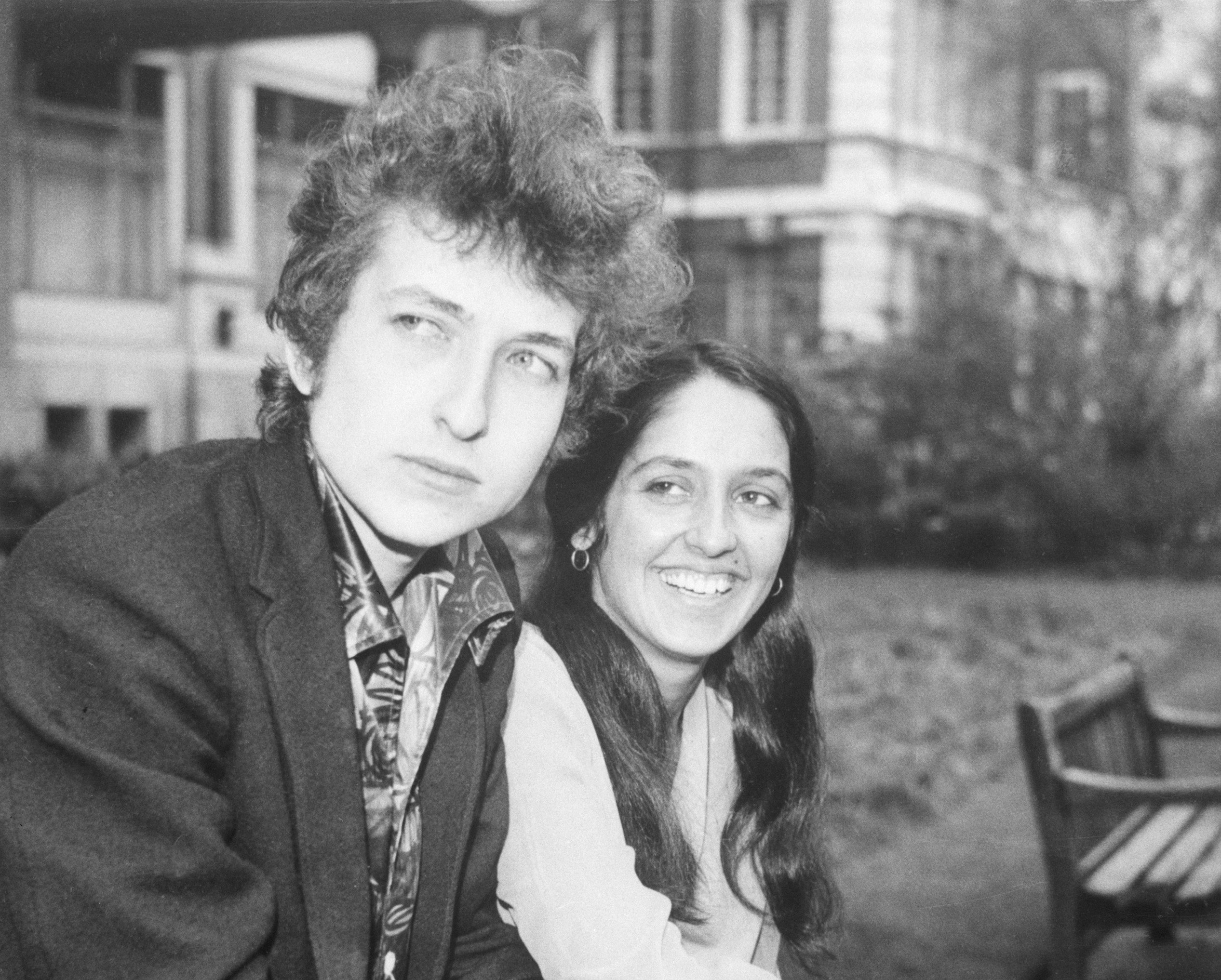 A black and white picture of Bob Dylan and Joan Baez sitting on a bench together outdoors. 