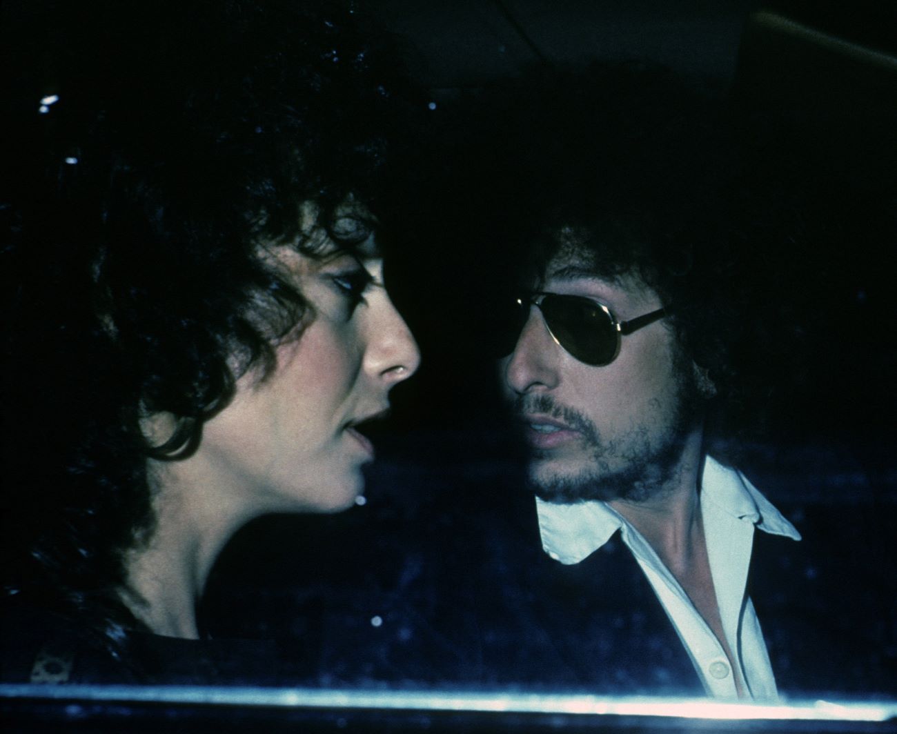 Sara Dylan and Bob Dylan sit in a car together. Bob Dylan wears sunglasses. 