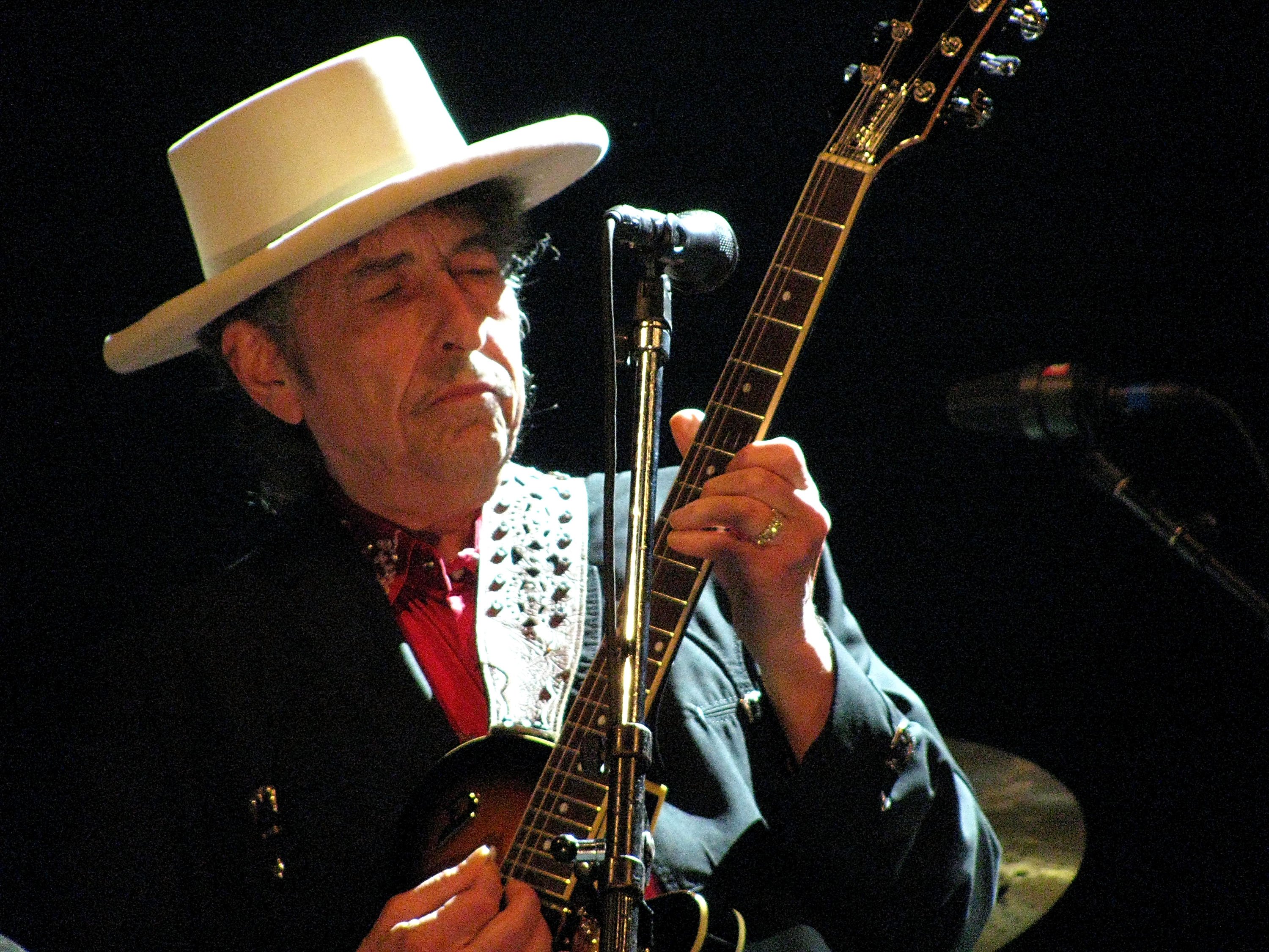 Bob Dylan wears a hat and plays the guitar in front of a microphone.