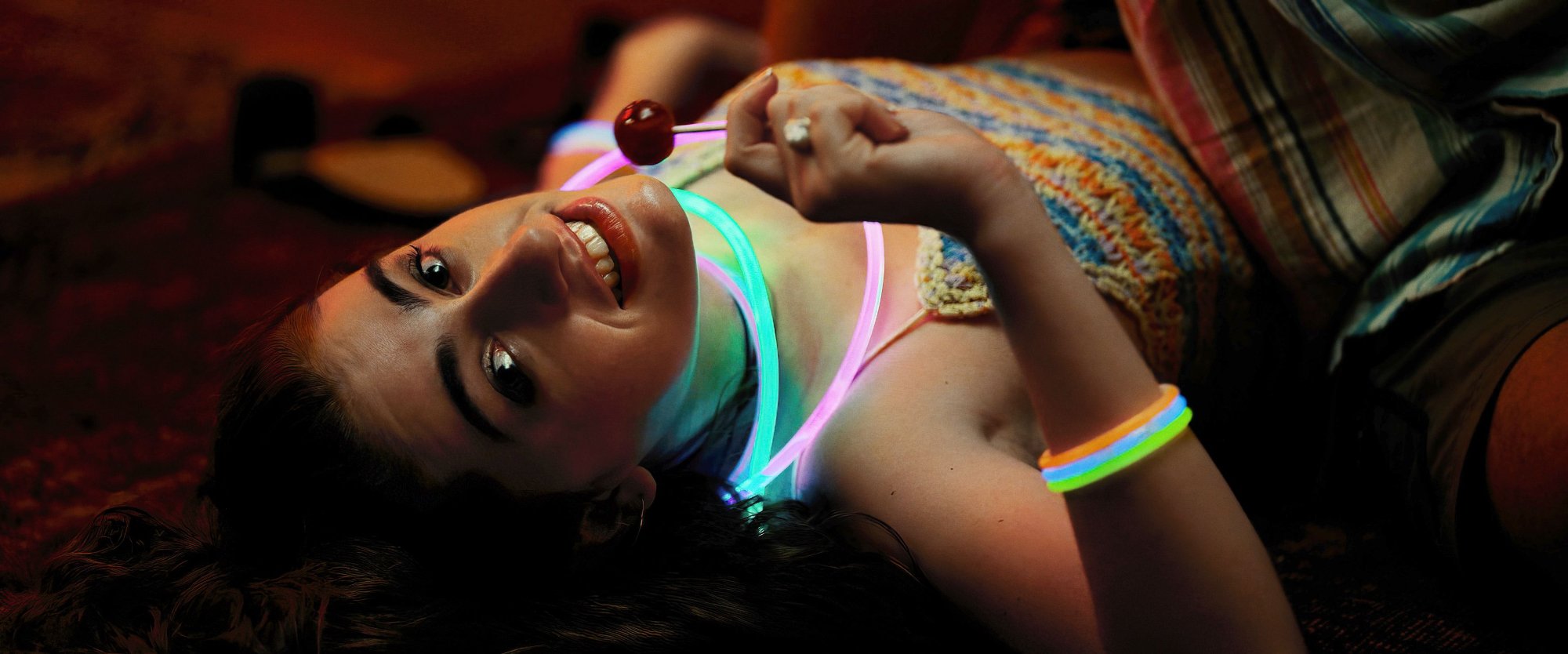 'Bodies Bodies Bodies' Rachel Sennott as Alice laying on her back with neon lights around her neck, smiling, holding a lollipop