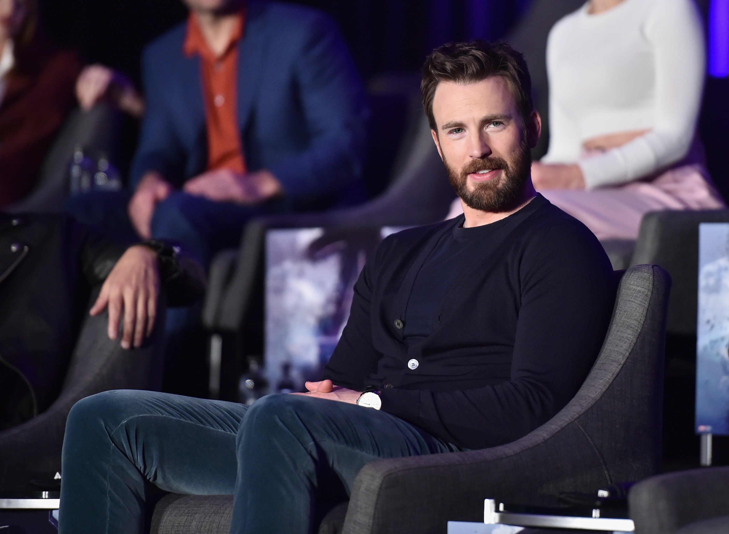 Chris Evans, who played Captain America in the MCU, wears a black cardigan over a dark blue shirt and blue velvet pants.