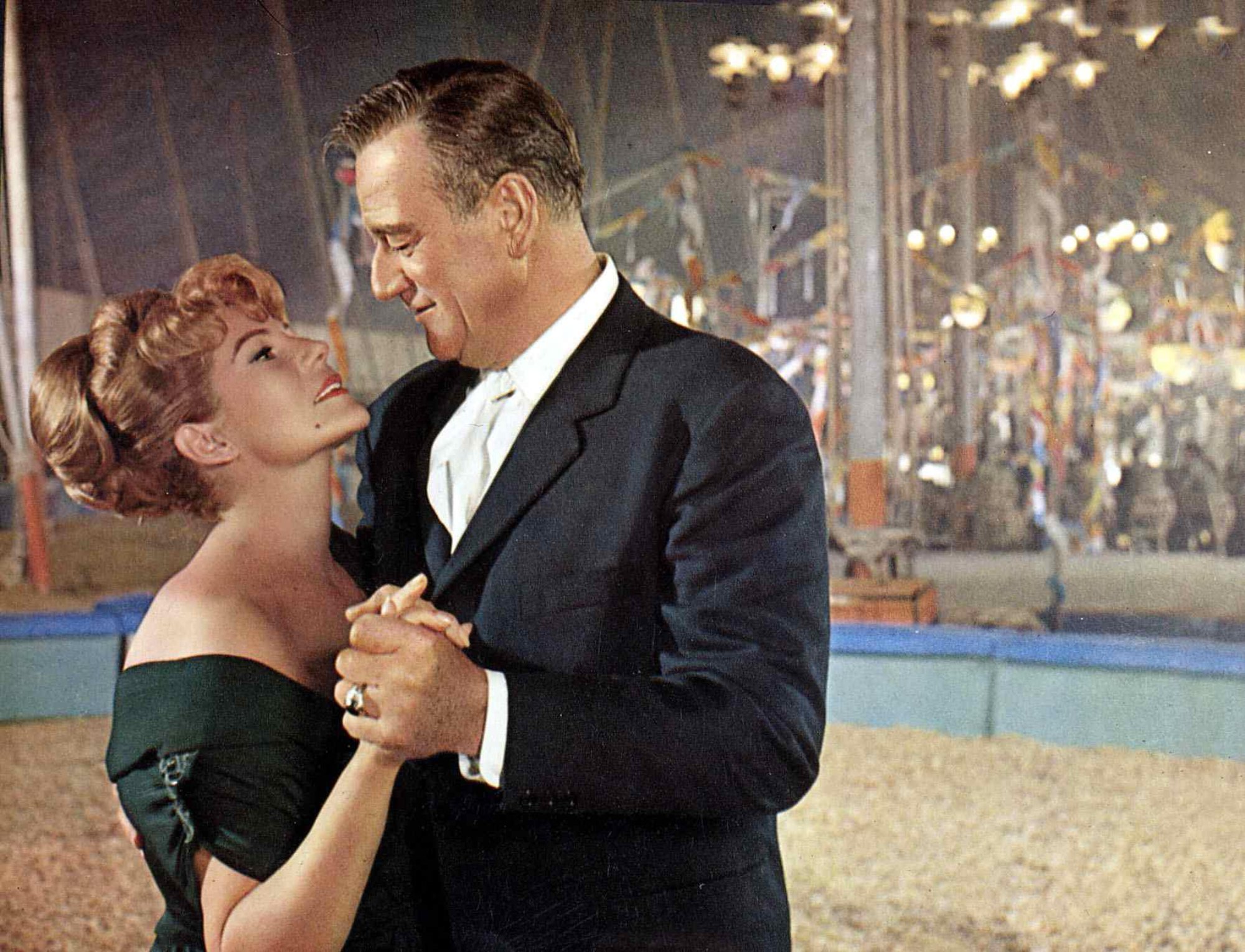 'Circus World' Rita Hayworth as Lili Alfredo and John Wayne as Matt Masters holding hands, looking into each other's eyes in front of circus