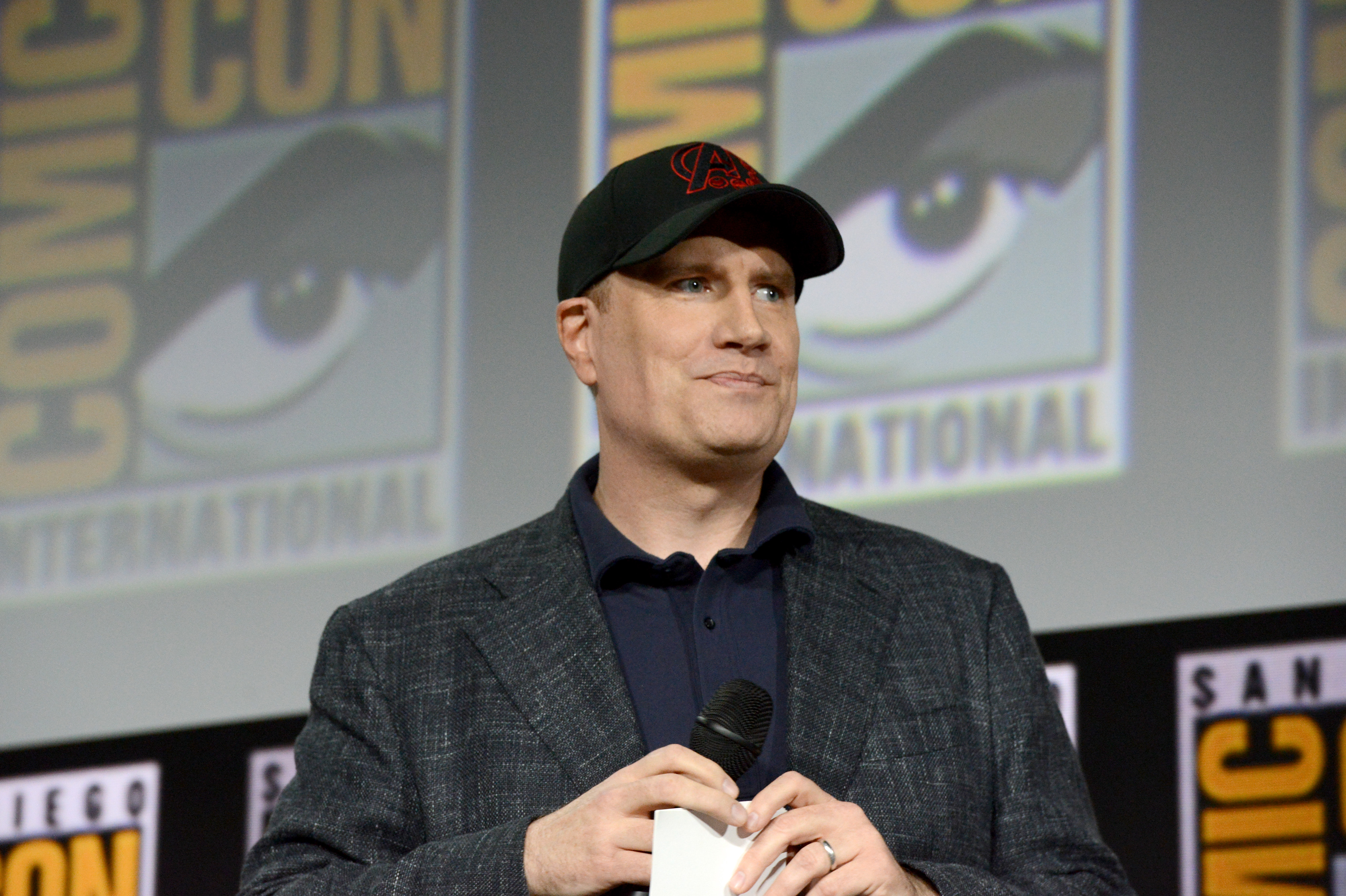 Marvel Studios president Kevin Feige appears onstage at San Diego Comic-Con in 2019. He wears a dark gray suit over a blue button-up shirt and a black baseball cap with a red Avengers logo on it.