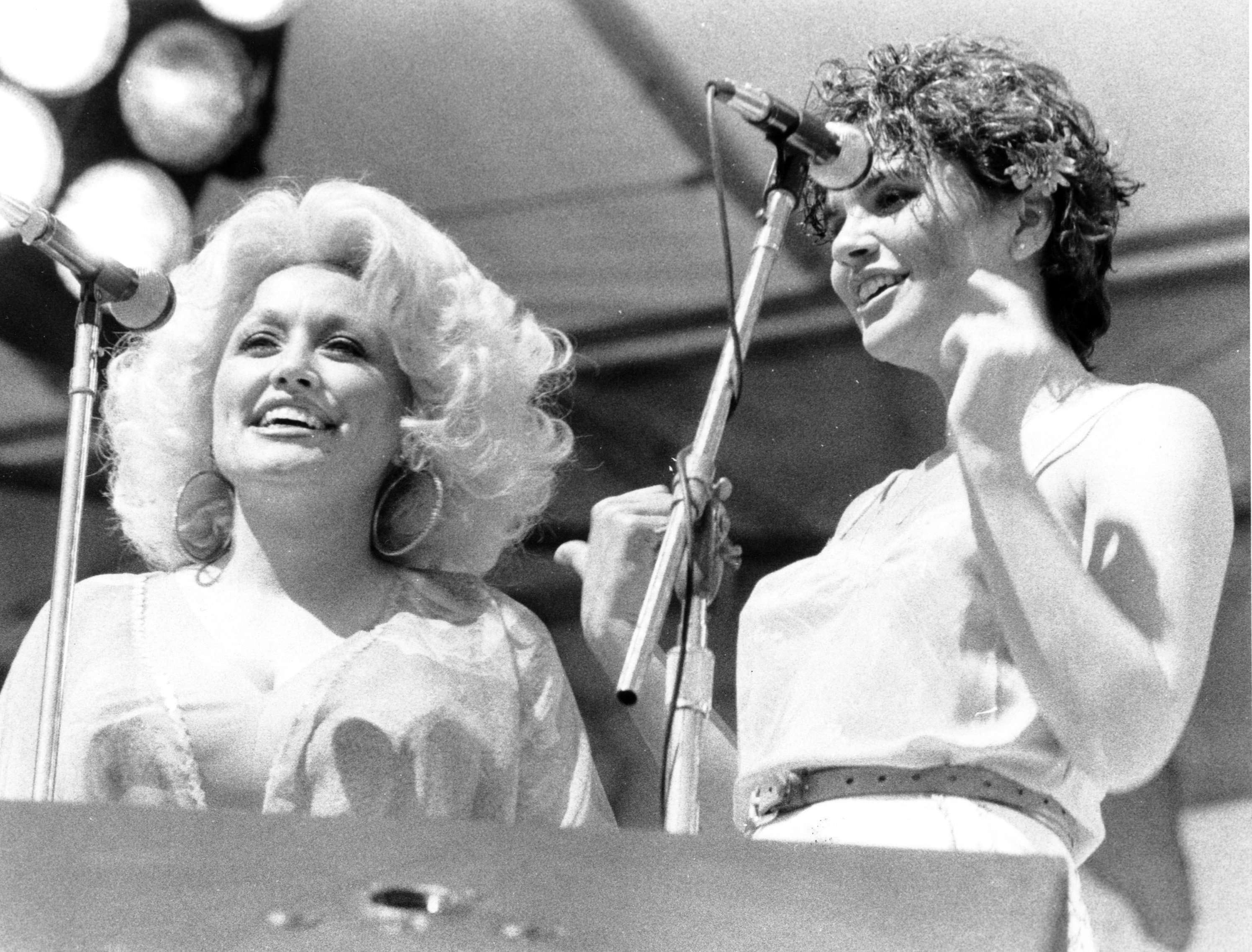 A black and white picture of Dolly Parton and Linda Ronstadt standing outside in front of two microphones.