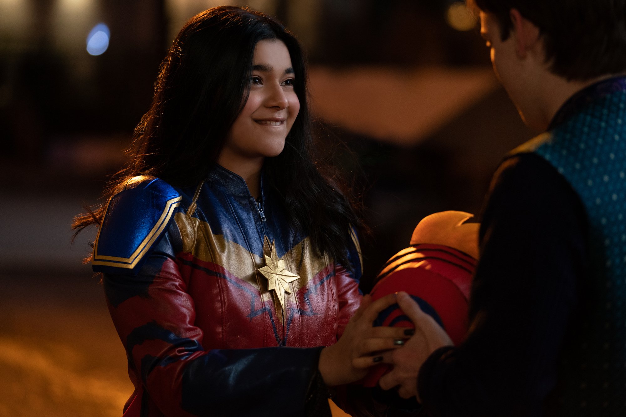 Iman Vellani as Kamala Khan in 'Ms. Marvel,' which fans want greenlit for season 2. She's wearing a red, blue, and gold suit, holding a helmet, and smiling at Bruno (Matt Lintz).
