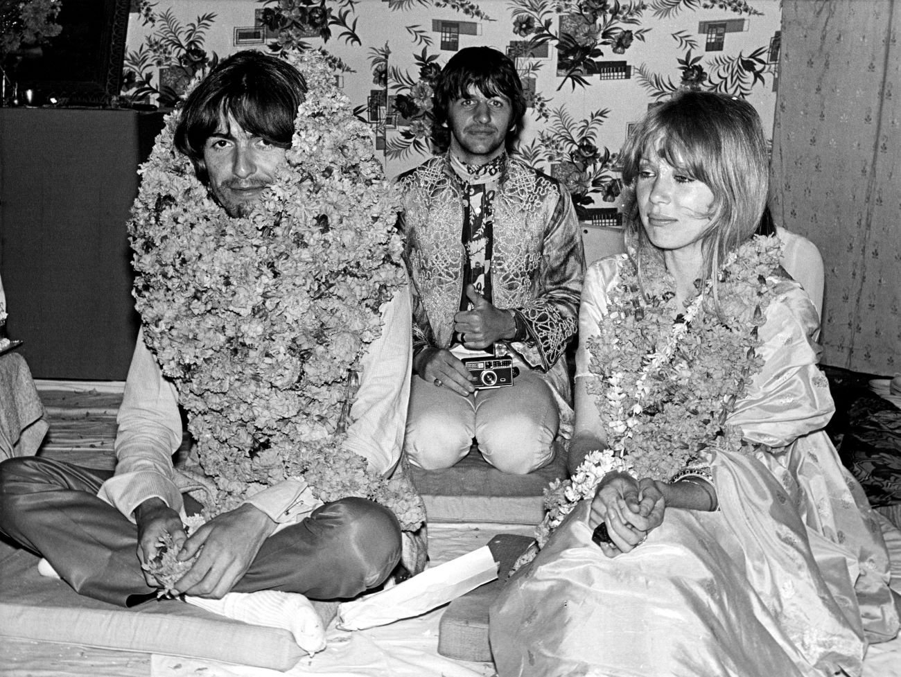 A black and white picture of George Harrison, Ringo Starr, and Pattie Boyd sitting on cushions with flowers around their necks. 