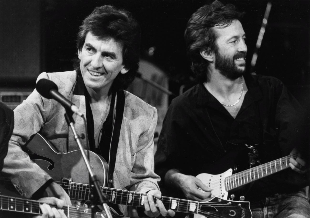 A black and white picture of George Harrison and Eric Clapton holding guitars and standing near a microphone.
