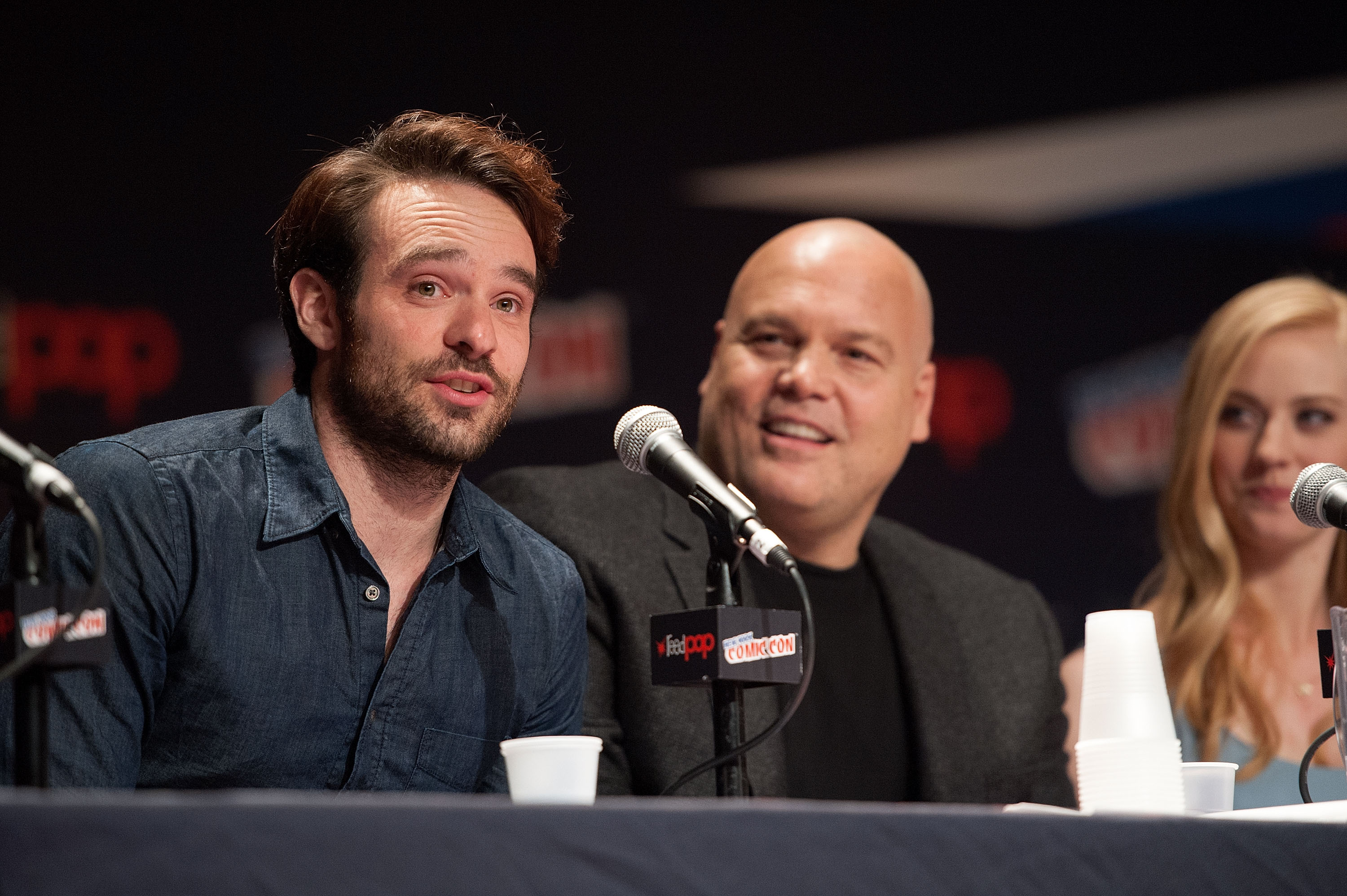 Charlie Cox, Vincent D'Onofrio, and Deborah Ann Woll, stars of 'Daredevil,' appear onstage at a panel. Cox wears a dark blue button-up shirt. D'Onofrio wears a dark gray suit over a black shirt. Woll wears a blue top.