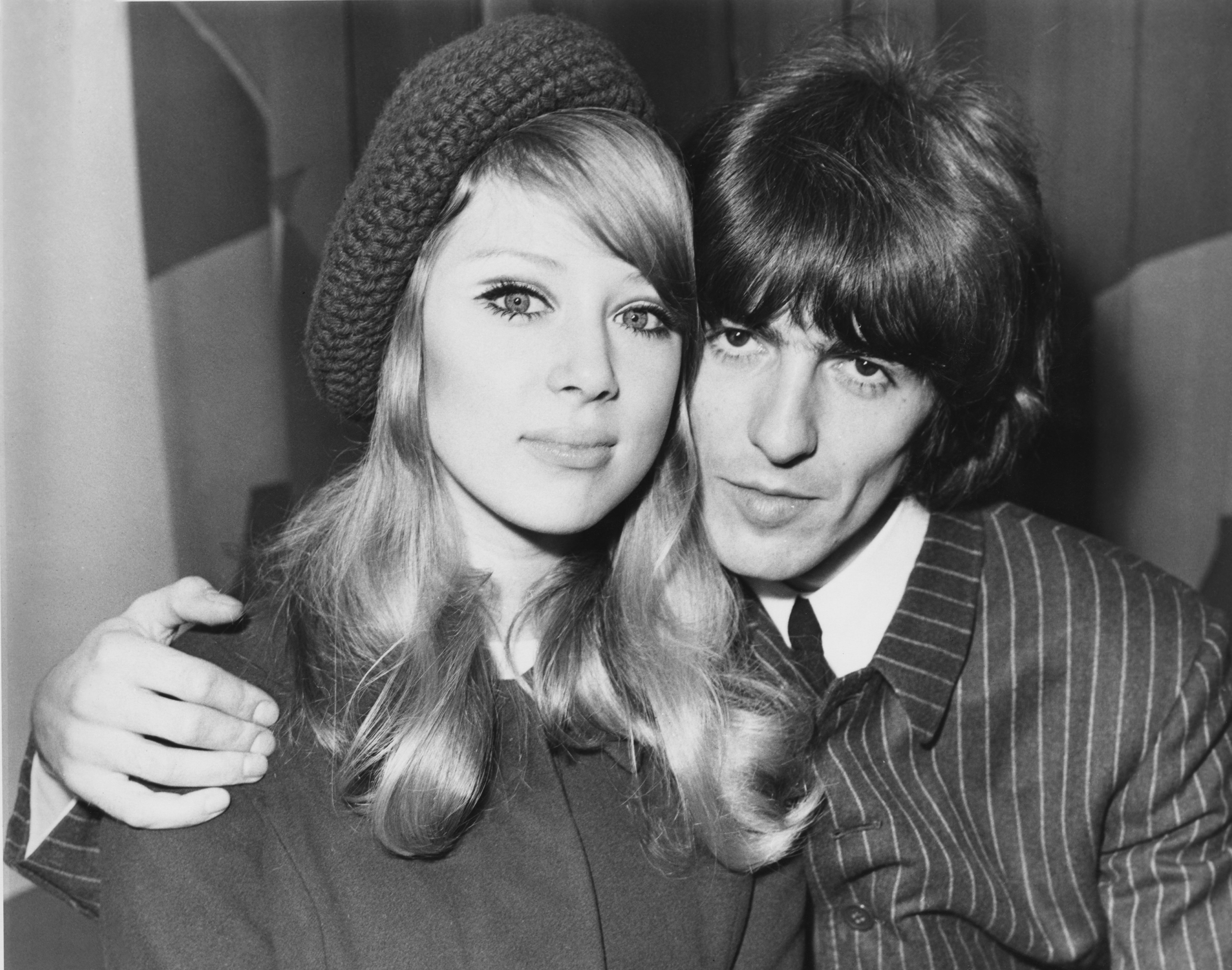 A black and white picture of George Harrison sitting with his arm around Pattie Boyd. She wears a knit hat.