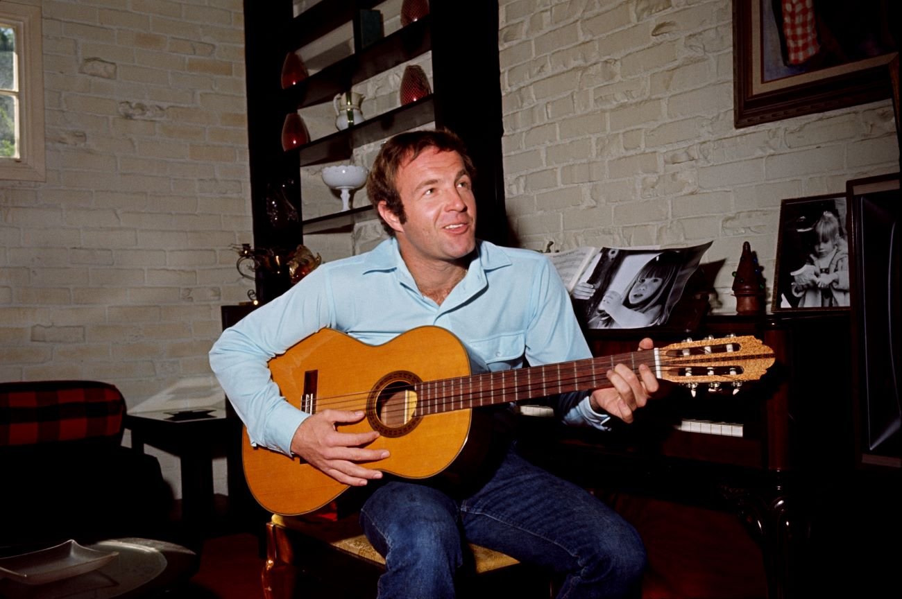 James Caan sits on a piano bench and holds a guitar.