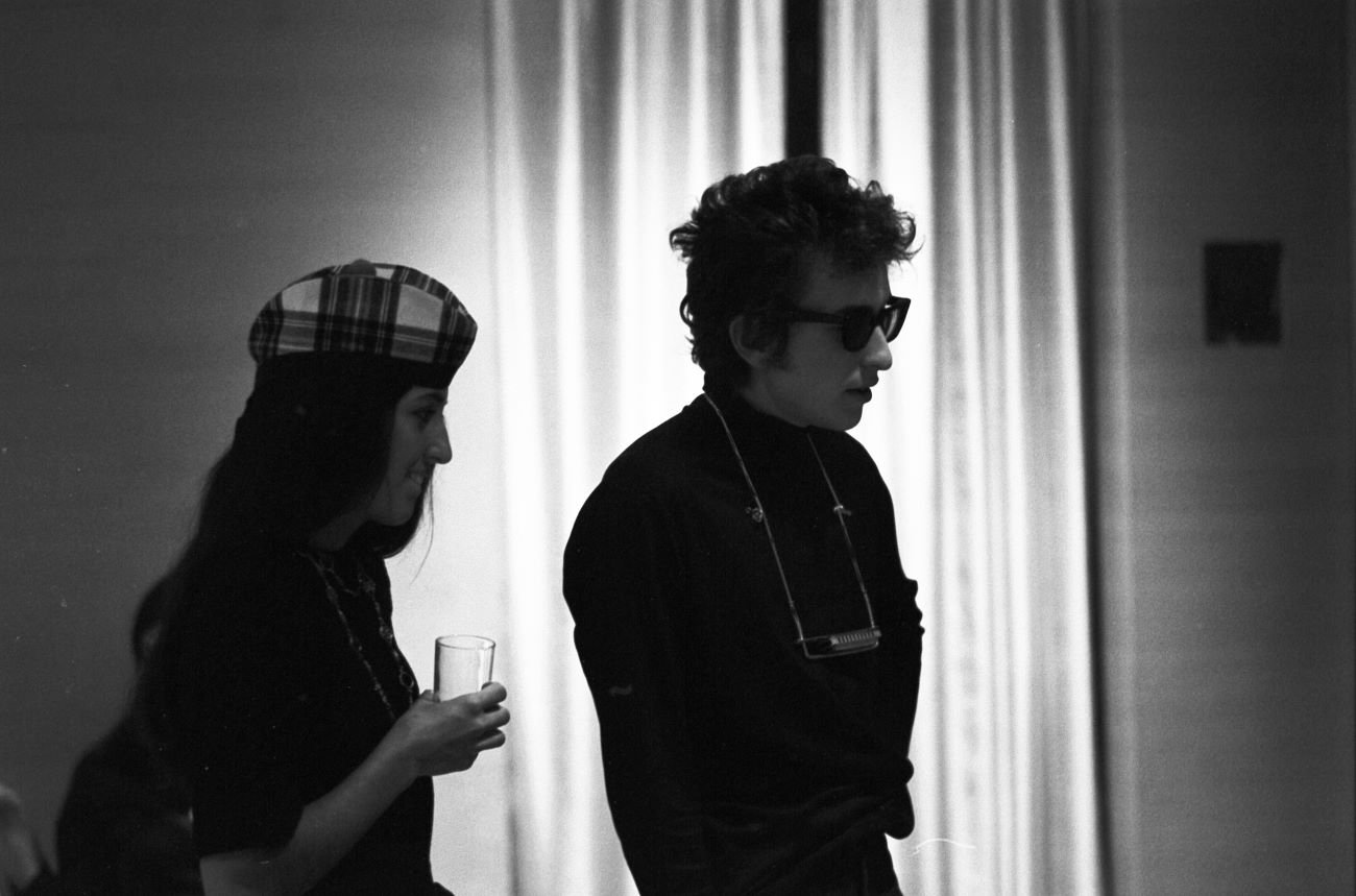 A black and white picture of Joan Baez wearing a hat and holding a class. Bob Dylan wears a pair of sunglasses