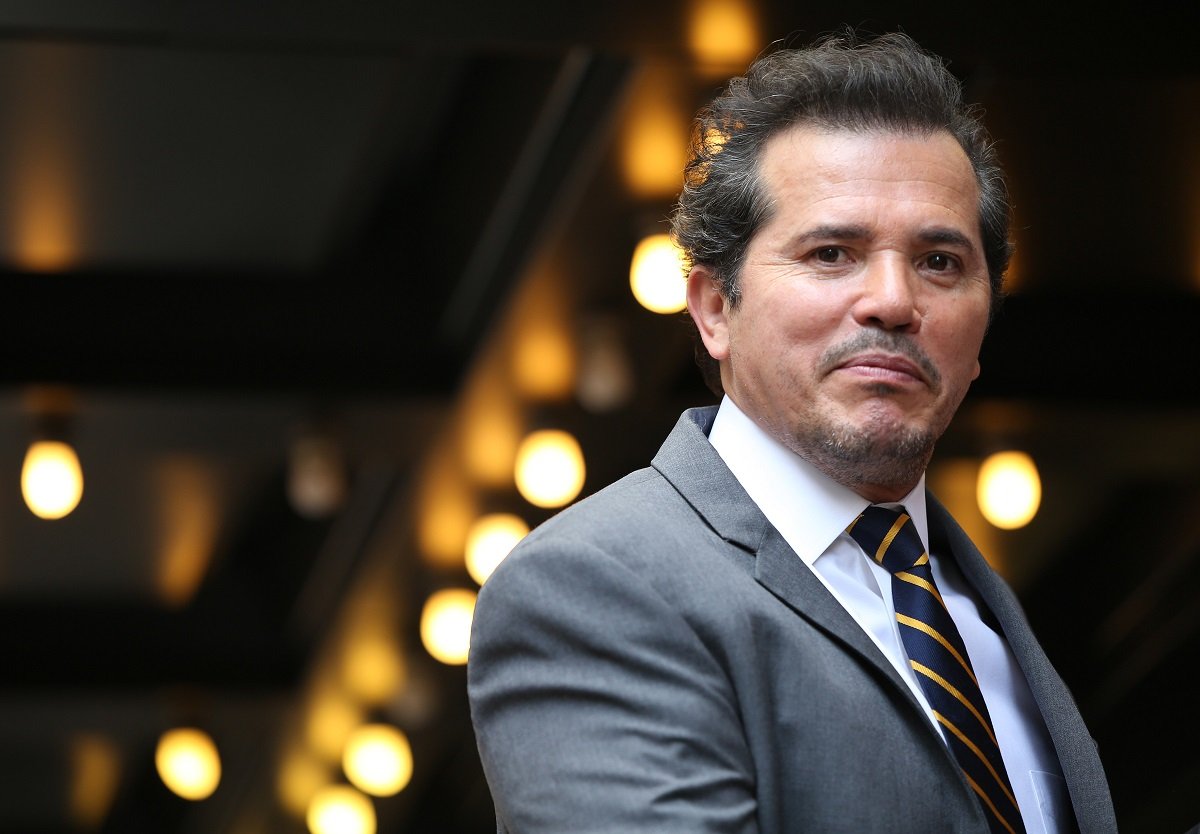 John Leguizamo smirking while wearing a grey suit.