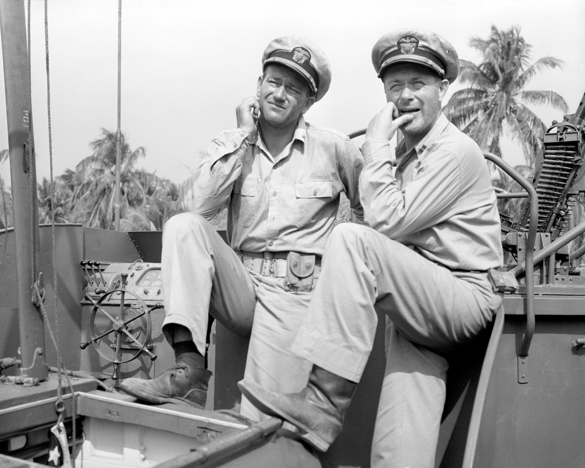 John Wayne as Lt. Rusty Ryan and Robert Montgomery as Lt. John Brickley, who had different career positions during World War II. They're wearing uniforms and squinting at the oncoming light while standing on a boat.