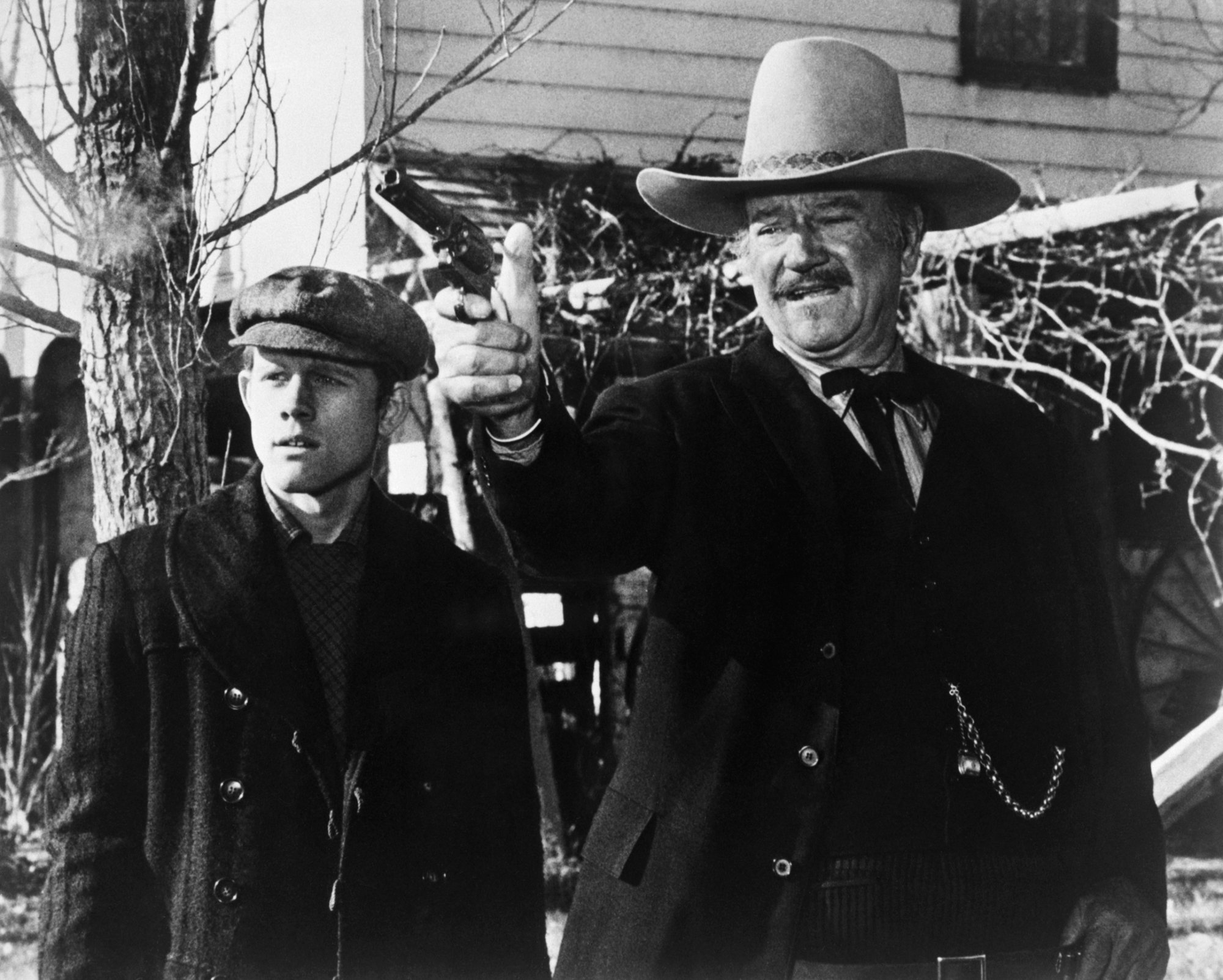 John Wayne in one of his last movies 'The Shootist' alongside Ron Howard. He's wearing a Western outfit and holding a gun, pointing it out standing next to a stunned Howard.