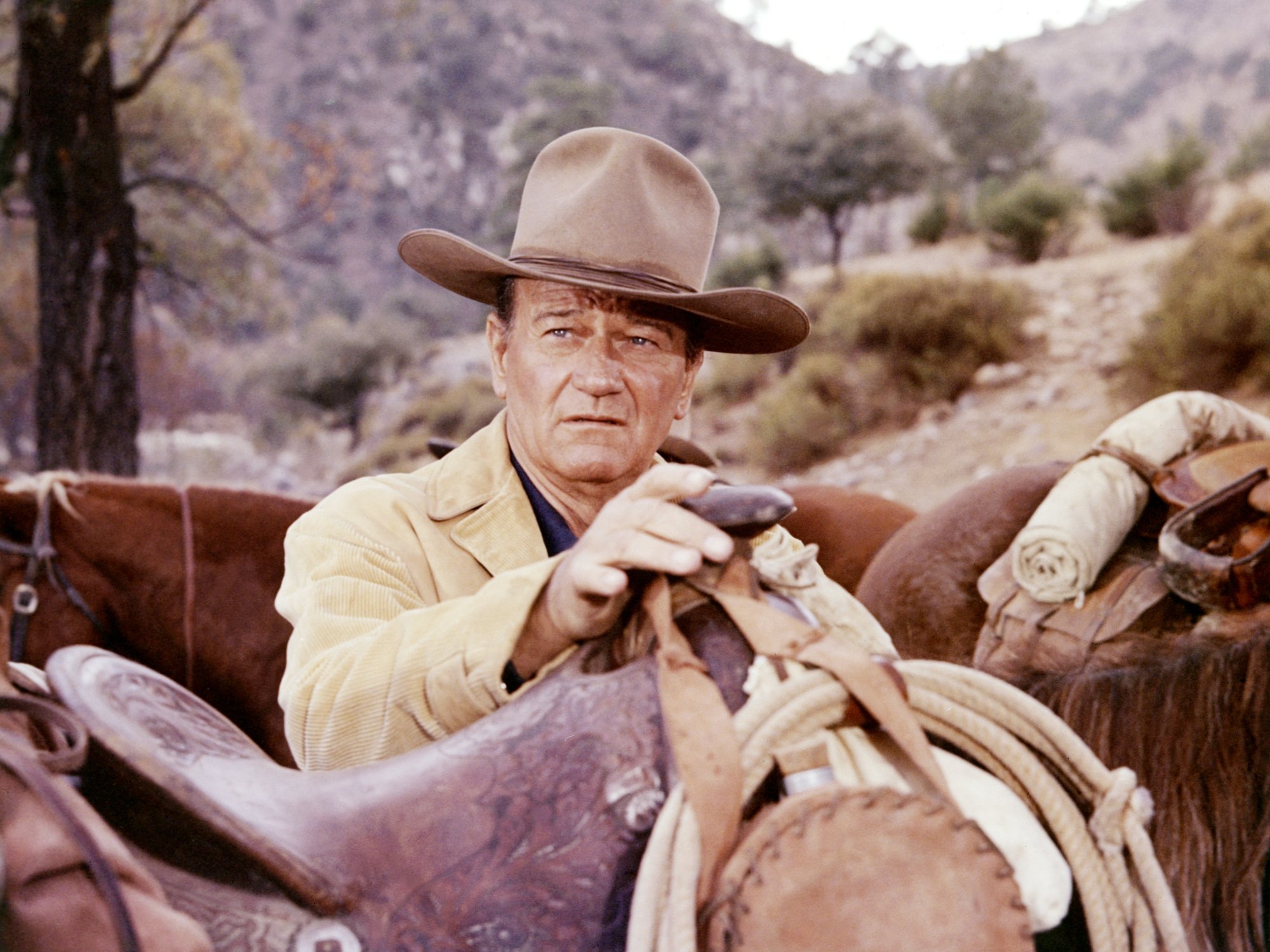 John Wayne movie star with his hand on the saddle of a horse while wearing a cowboy uniform