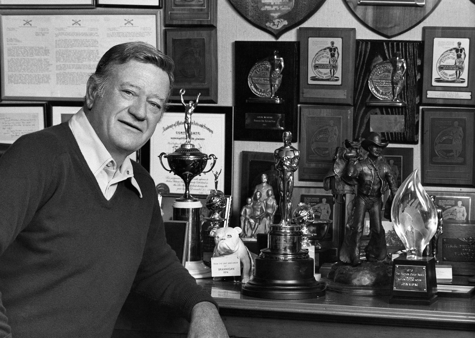 John Wayne, who starred in Western and war movies, smiling in front of a wall of trophies and plaques