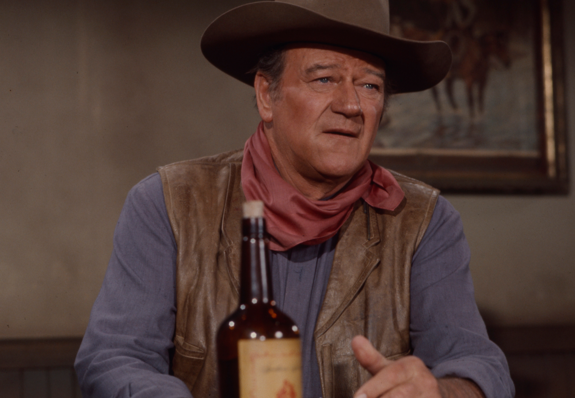 John Wayne, who starred in Western movies, wearing a cowboy costume and hat with a bottle in front of him while filming 'Rio Lobo'
