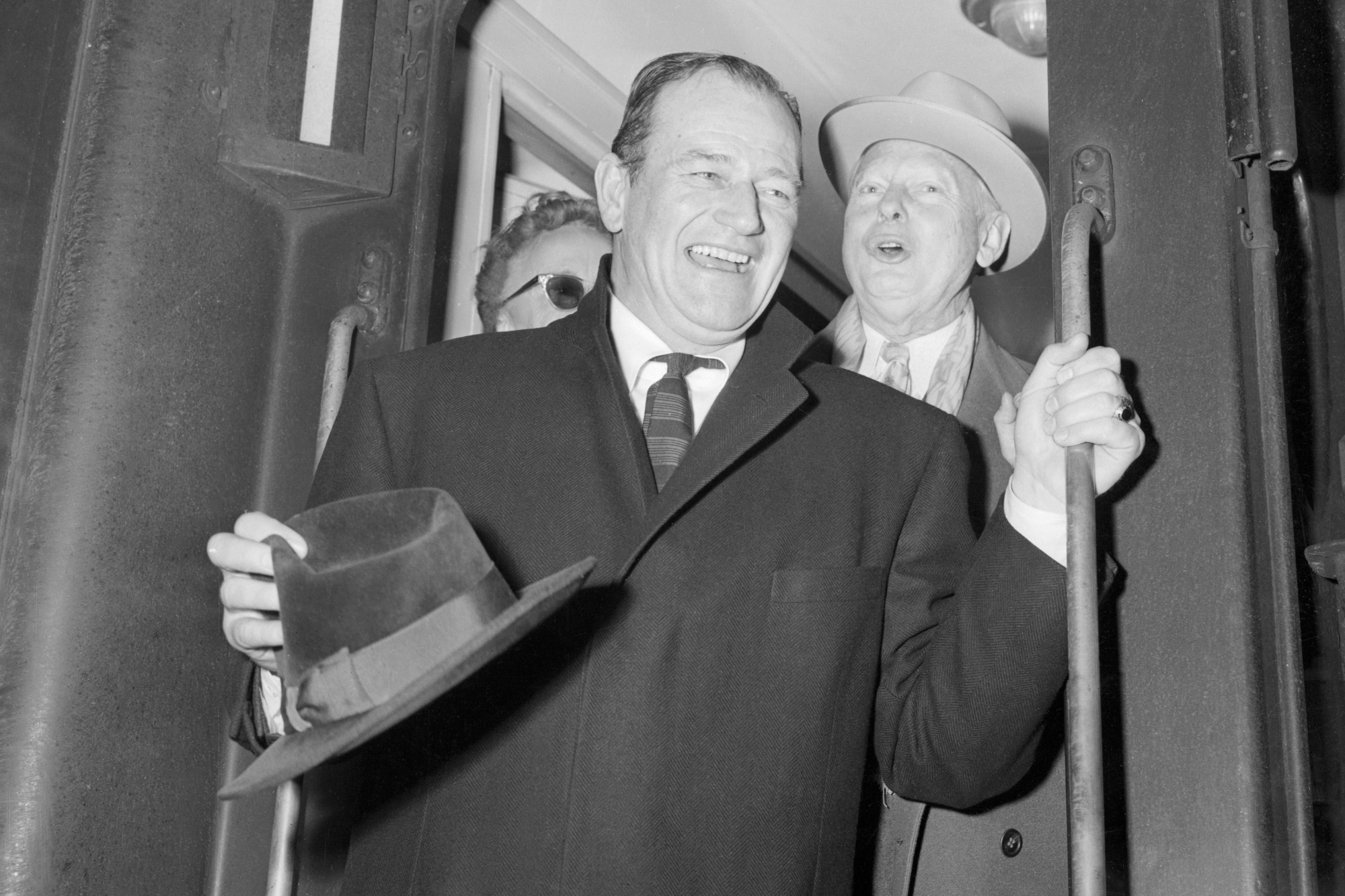John Wayne, who starred in the movie 'The Conqueror,' smiling holding a hat, while wearing a suit