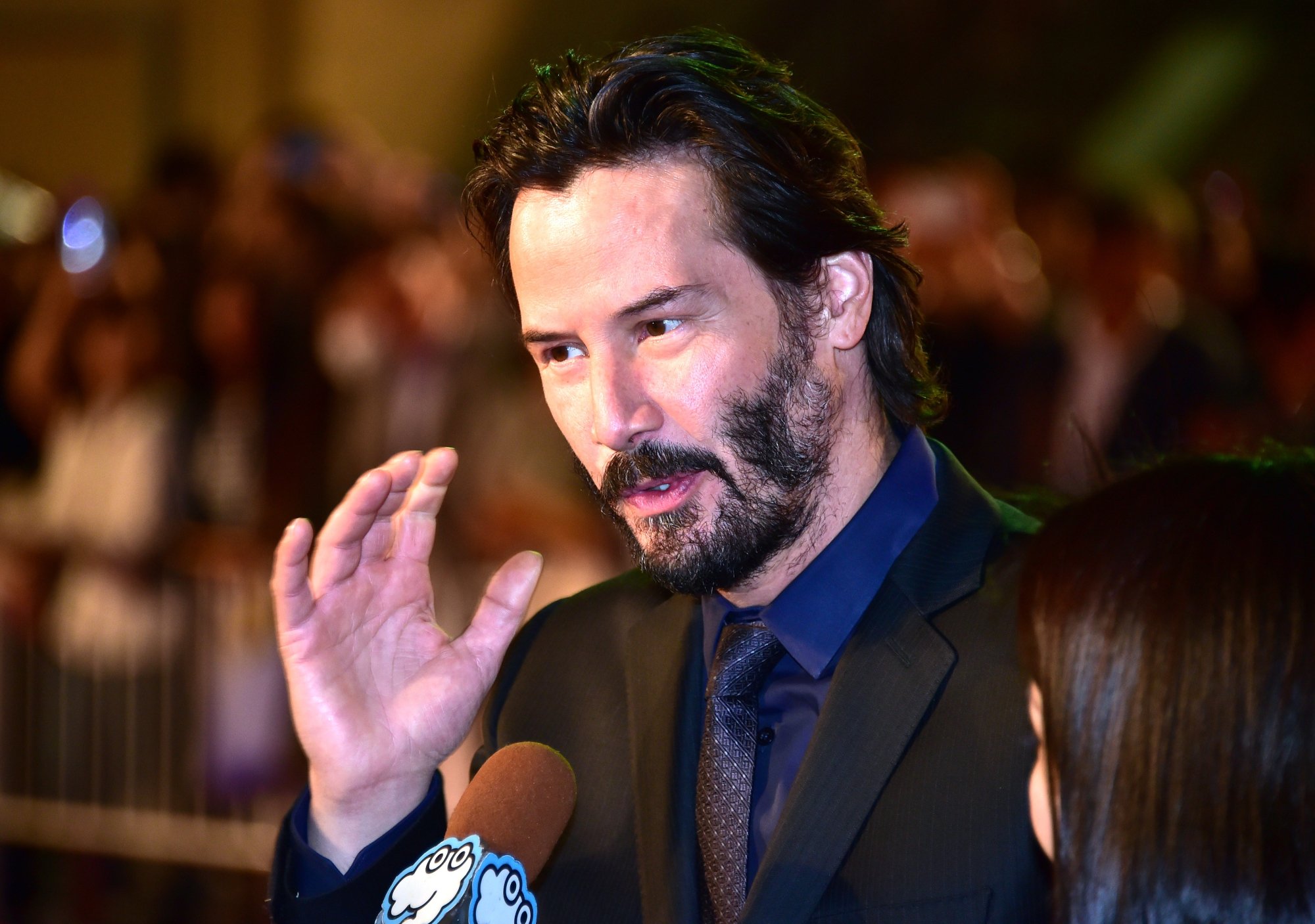 'John Wick' actor Keanu Reeves wearing a black suit holding his hand up while talking