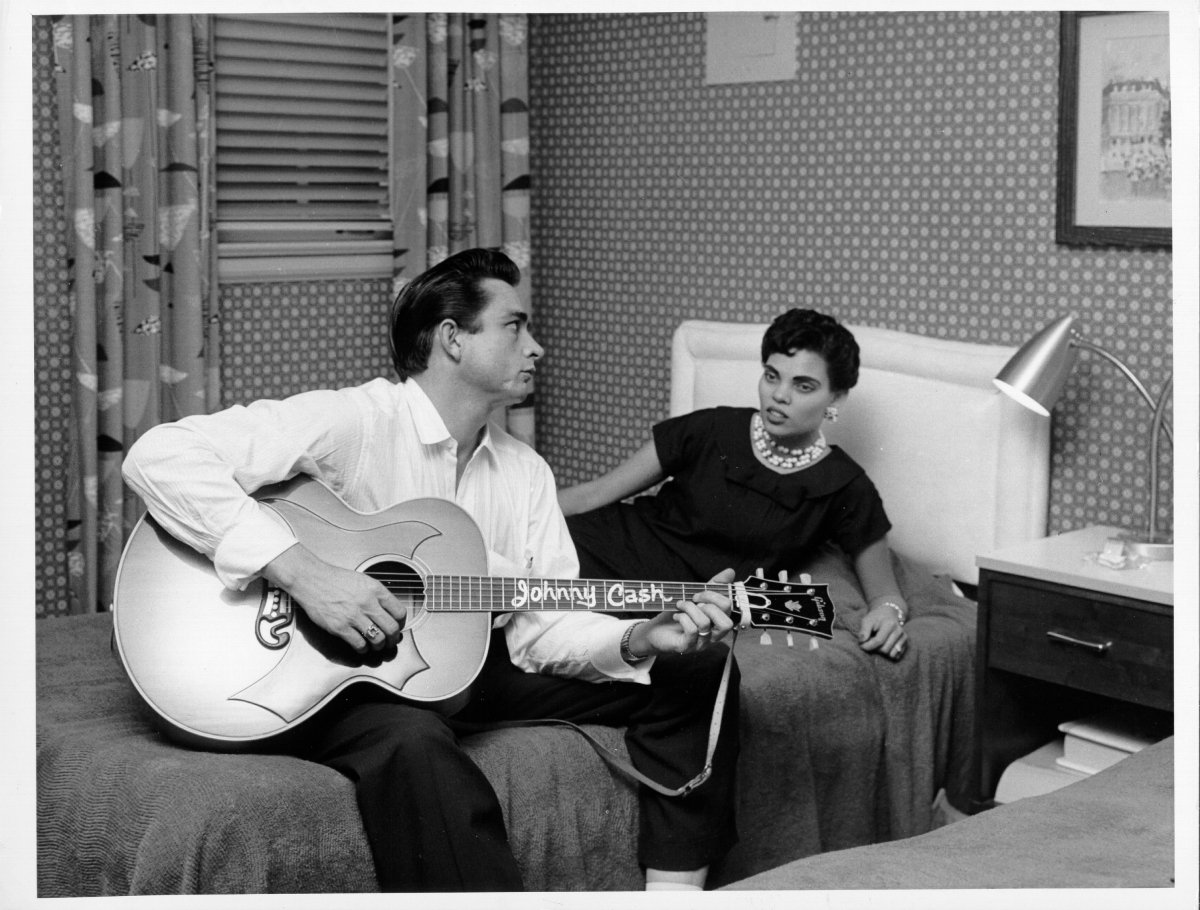 Johnny Cash sits on a bed playing acoustic guitar as his first wife Vivian Liberto looks on in circa 1957