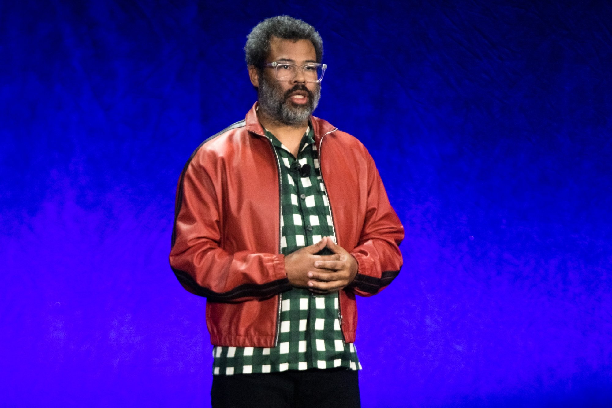 Jordan Peele 'Nope' wearing an red jacket with his hands crossed in front of him while standing in front of a blue background
