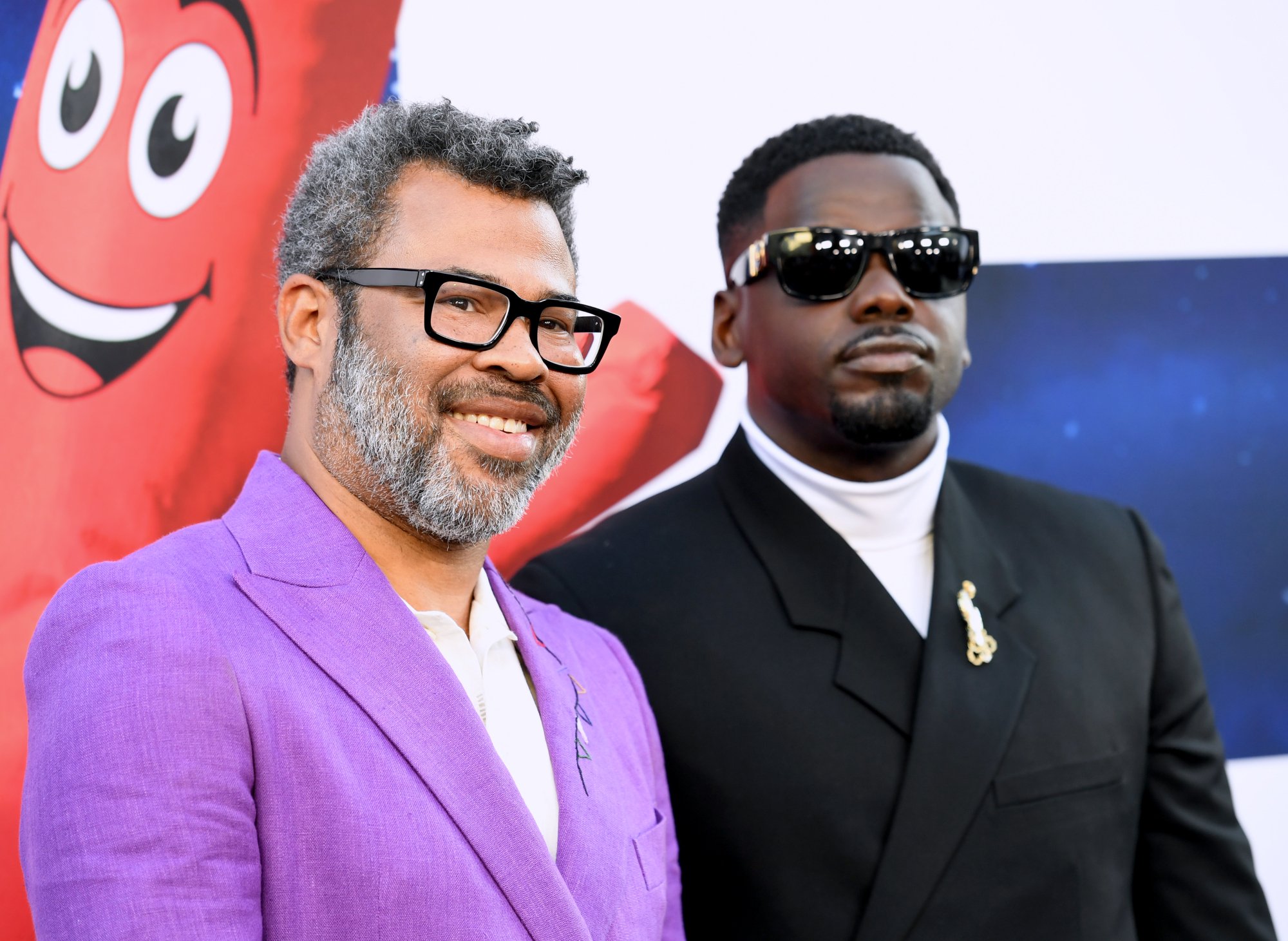 Jordan Peele and Daniel Kaluuya standing next to each other in purple and black suits, respectively. Peele is smiling and Kaluuya has a straight face as they stand in front of a waving inflatable arms man.