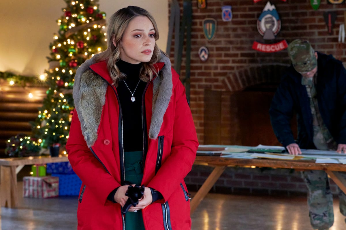 Kayla Wallace wearing a red coat and standing in front of a Christmas tree in the Hallmark Channel movie 'My Grown-Up Christmas List'