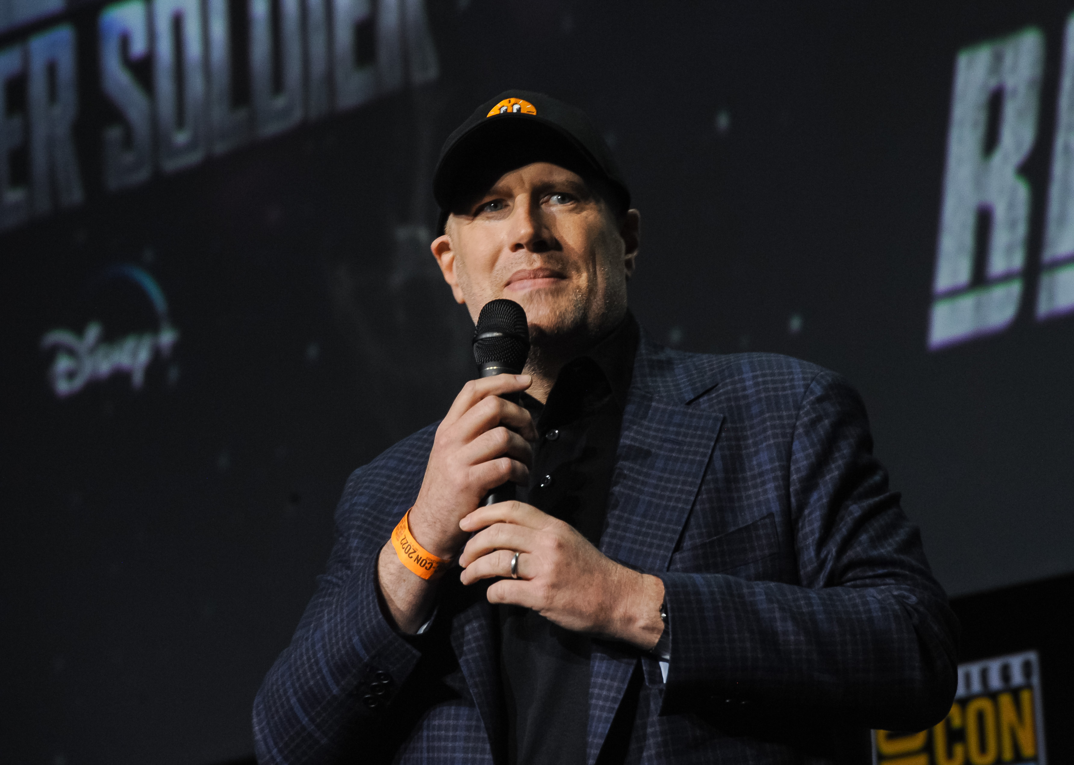 Kevin Feige, who spearheaded Phase 4 of the MCU, wears a dark plaid suit over a black button-up shirt and a black baseball cap with an image of Miss Minutes from 'Loki' on the front.