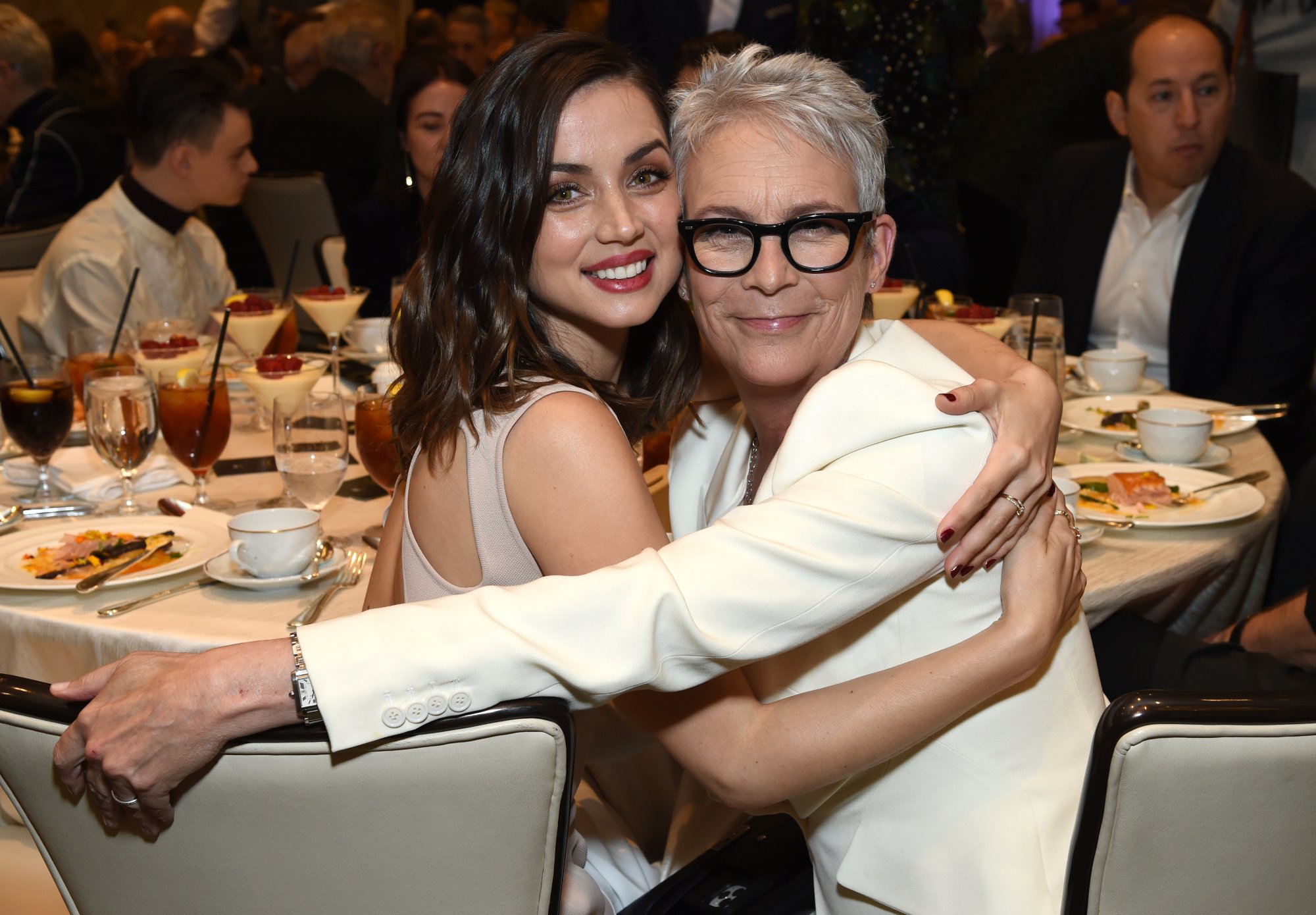 'Knives Out' actors Ana de Armas and Jamie Lee Curtis sitting next to each other at a table smiling, hugging each other.