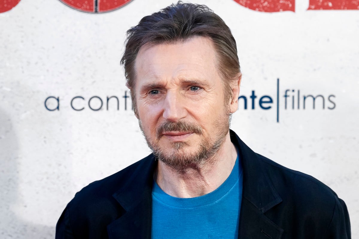 Liam Neeson posing while wearing a black jacket and blue shirt.