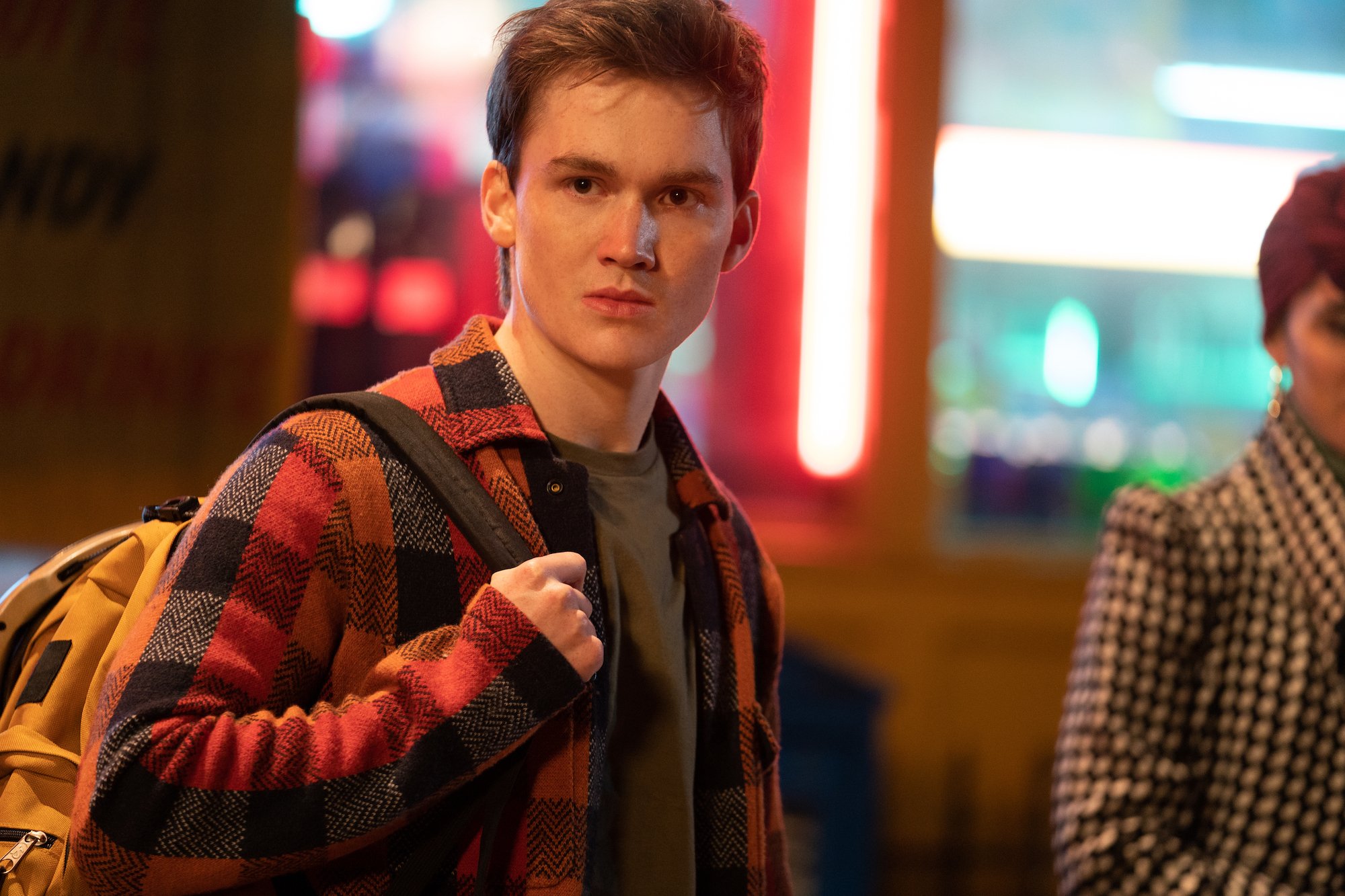 Matt Lintz, in character as Bruno in 'Ms. Marvel' on Disney+, wears a red and black plaid button-up shirt over a gray t-shirt.