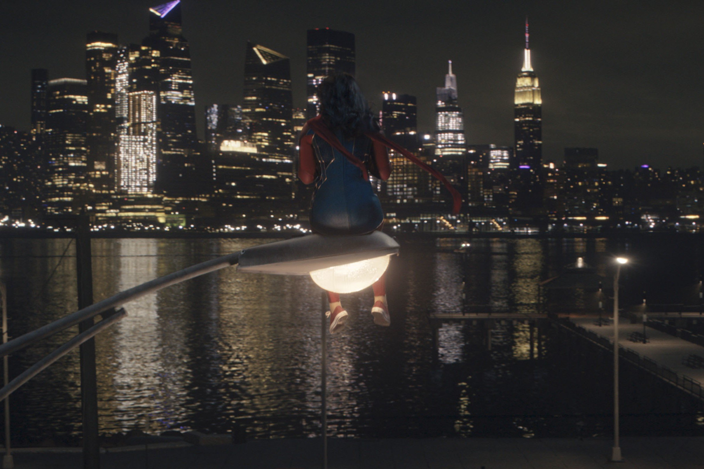 Iman Vellani, in character as Kamala Khan in 'Ms. Marvel,' which might have a season 2 on Disney+, wears her Ms. Marvel costume and sits on a street light and looks at the New York City skyline.