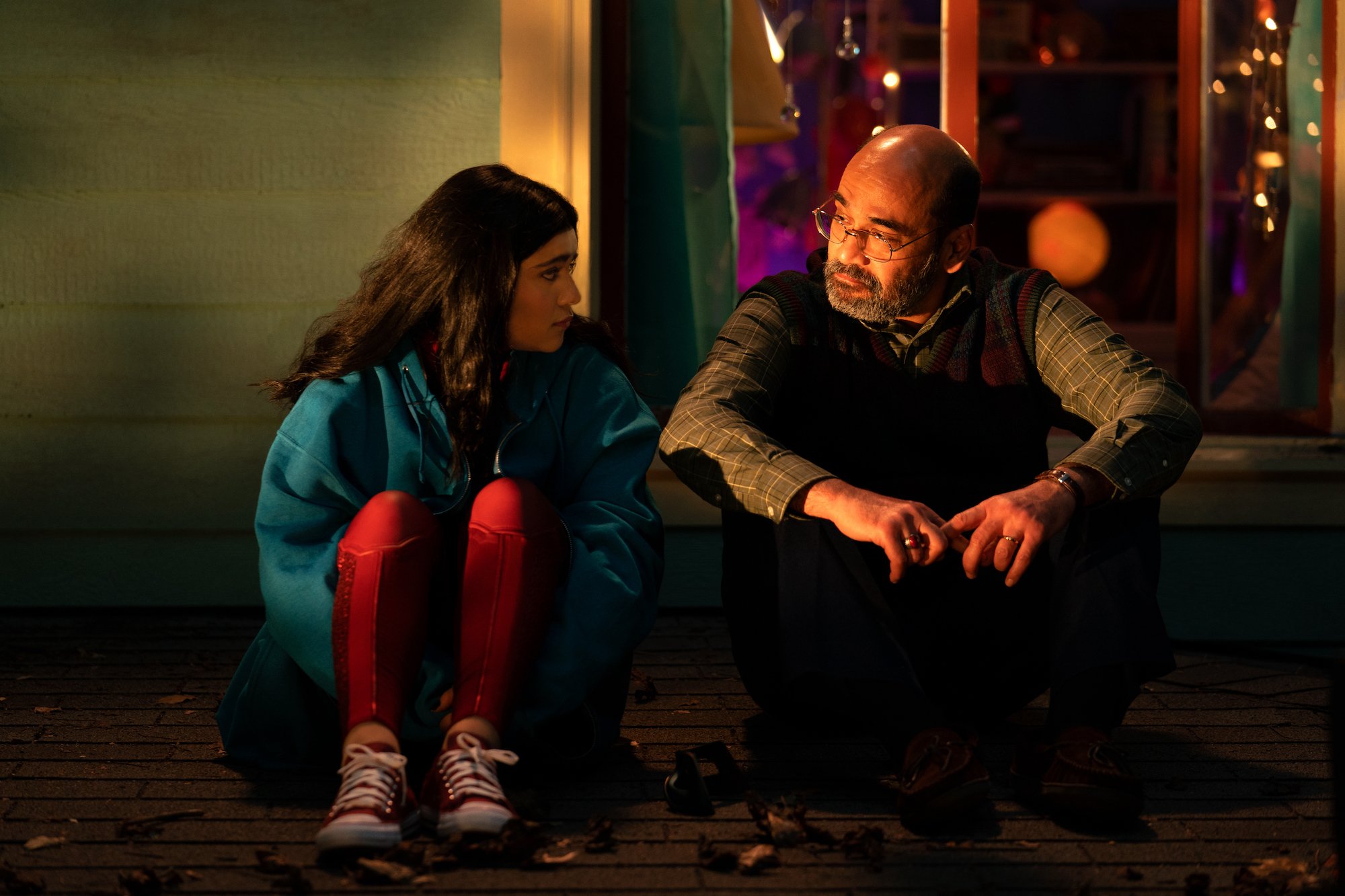 Iman Vellani and Mohan Kapur, in character as Kamala and Yusuf Khan, share a rooftop scene in 'Ms. Marvel' Episode 6 before the post-credits. Kamala wears a light blue jacket over her Ms. Marvel costume. Yusuf wears a red and green plaid sweater vest over a green plaid button-up shirt.