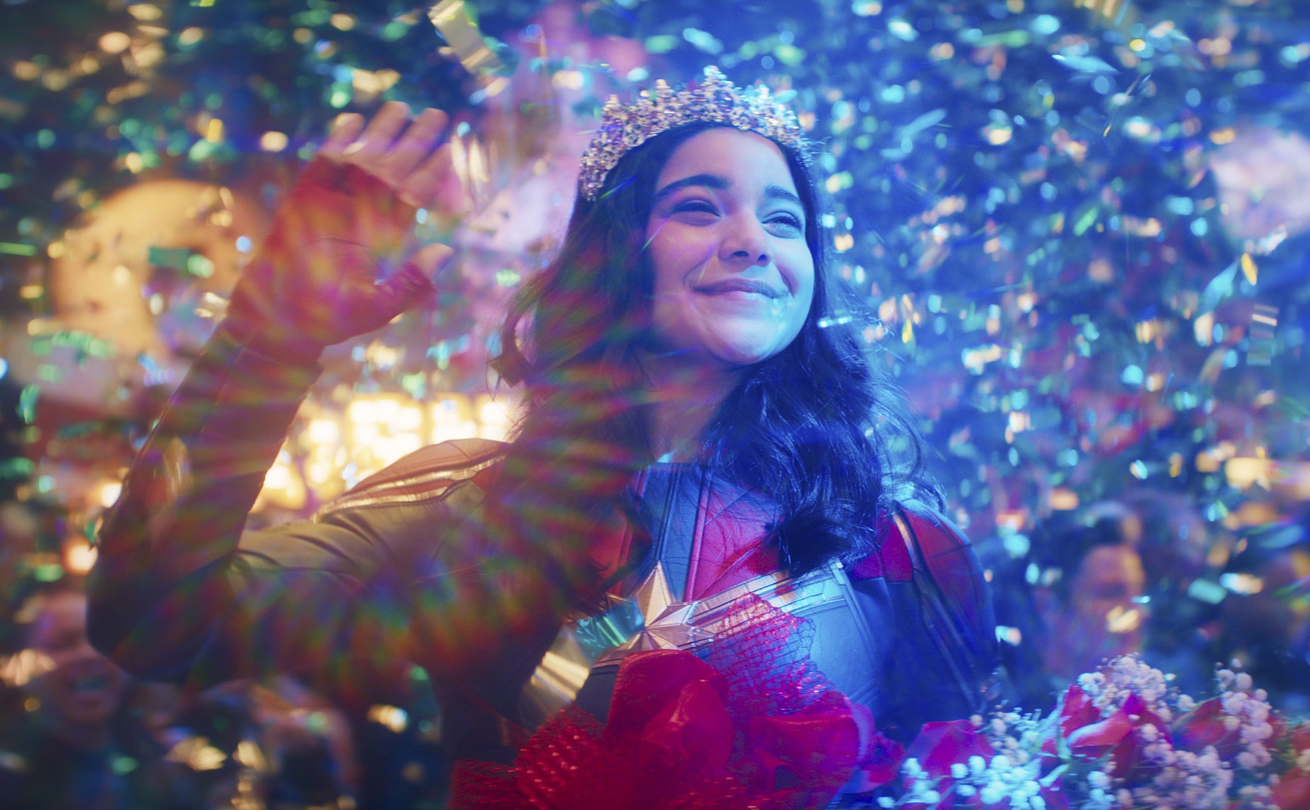 Iman Vellani as Kamala Khan in 'Ms. Marvel' Season 1. She's wearing a tiara, standing on stage, and waving at an audience.