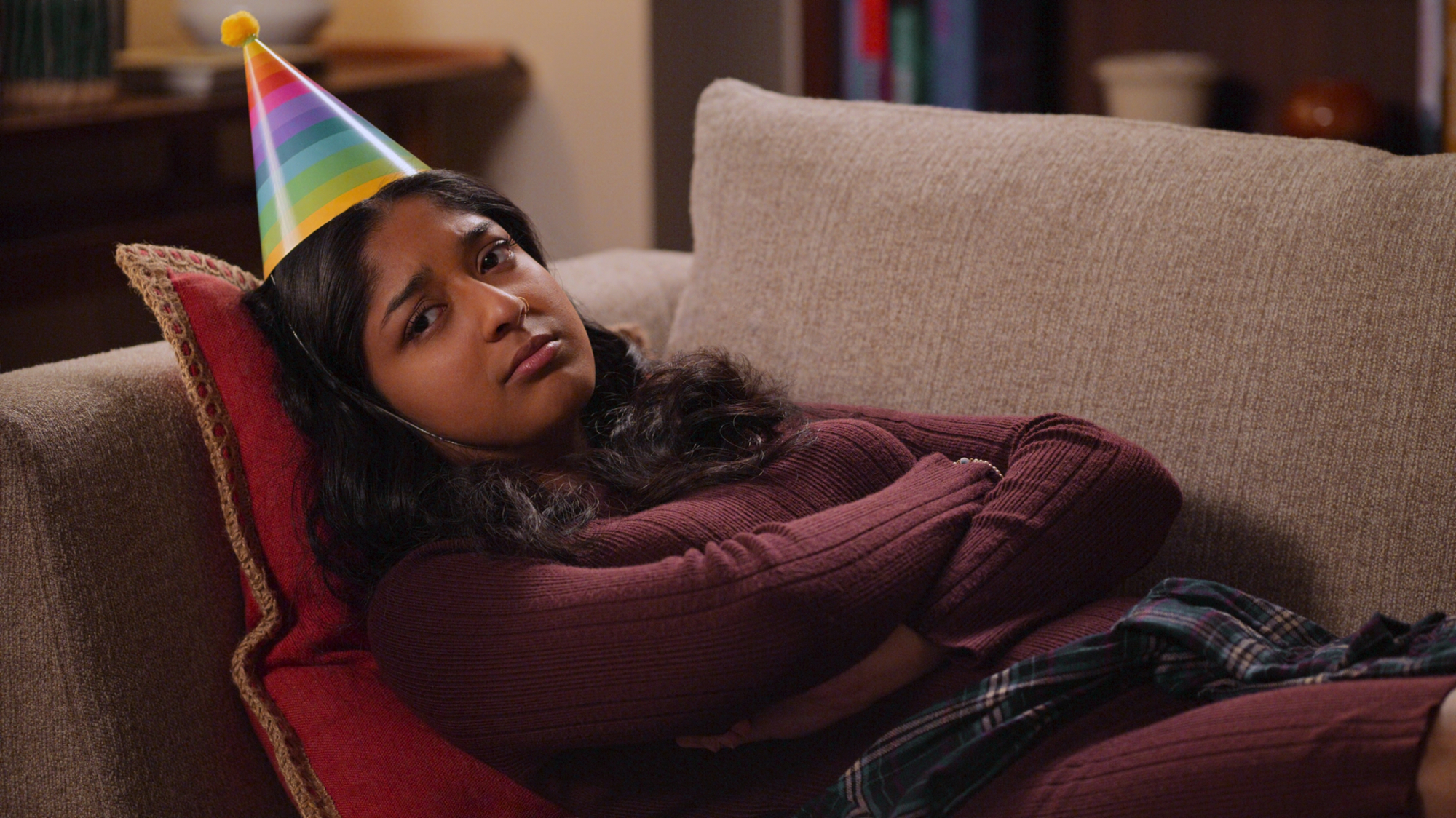 Maitreyi Ramakrishnan as Devi in 'Never Have I Ever,' a show that is on our list of series ending next season. She's lying on a couch with a birthday hat on and pouting.