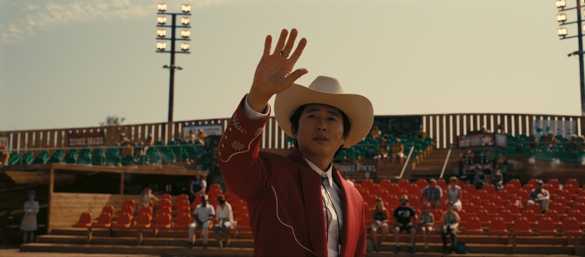 'Nope' Steven Yeun as Ricky 'Jupe' Park holding his hand up wearing a red suit jacket and cowboy hat in front of a crowd while holding his hand up