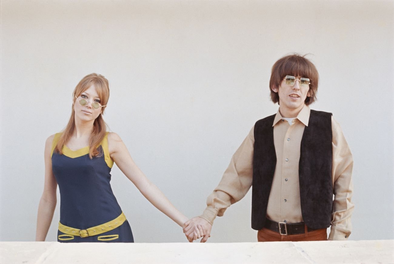 Pattie Boyd and George Harrison hold hands and stand in front of a white wall.