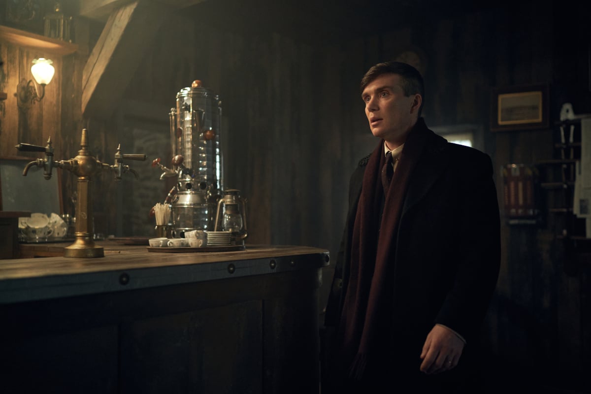 Peaky Blinders will be made into a movie. Thomas Shelby stands in a bar wearing a suit and red scarf. 