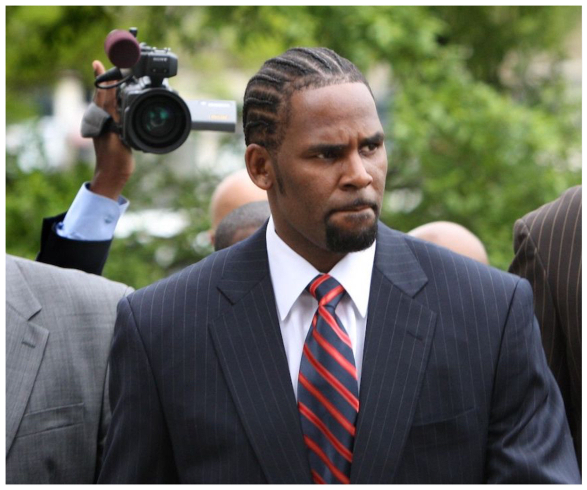 R. Kelly frowns and wears a suit as photographers snap his photo.
