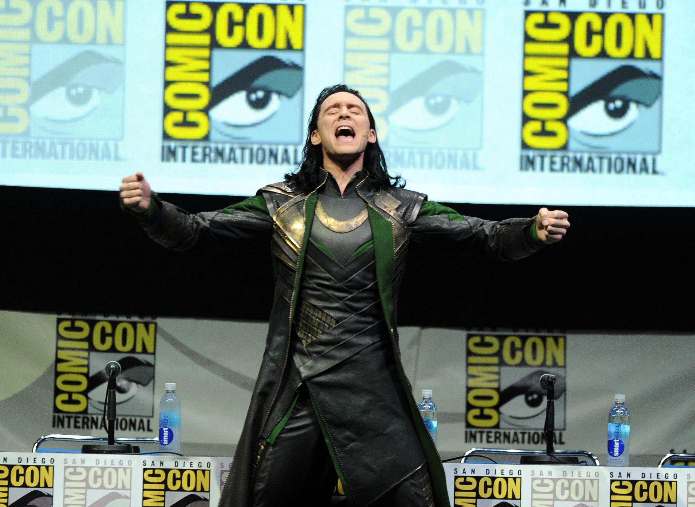 Tom Hiddleston as Loki stands on stage in front of a San Diego Comic Con poster