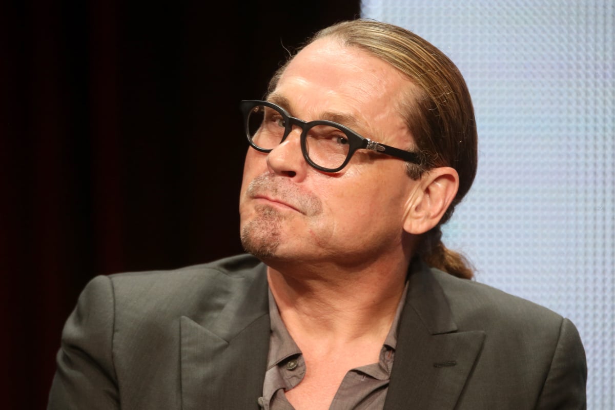 Kurt Sutter speaks onstage at the Sons of Anarchy panel during the FX Networks portion of the 2014 Summer Television Critics Association. Sutter wears a suit jacket, button-down shirt, and glasses.
